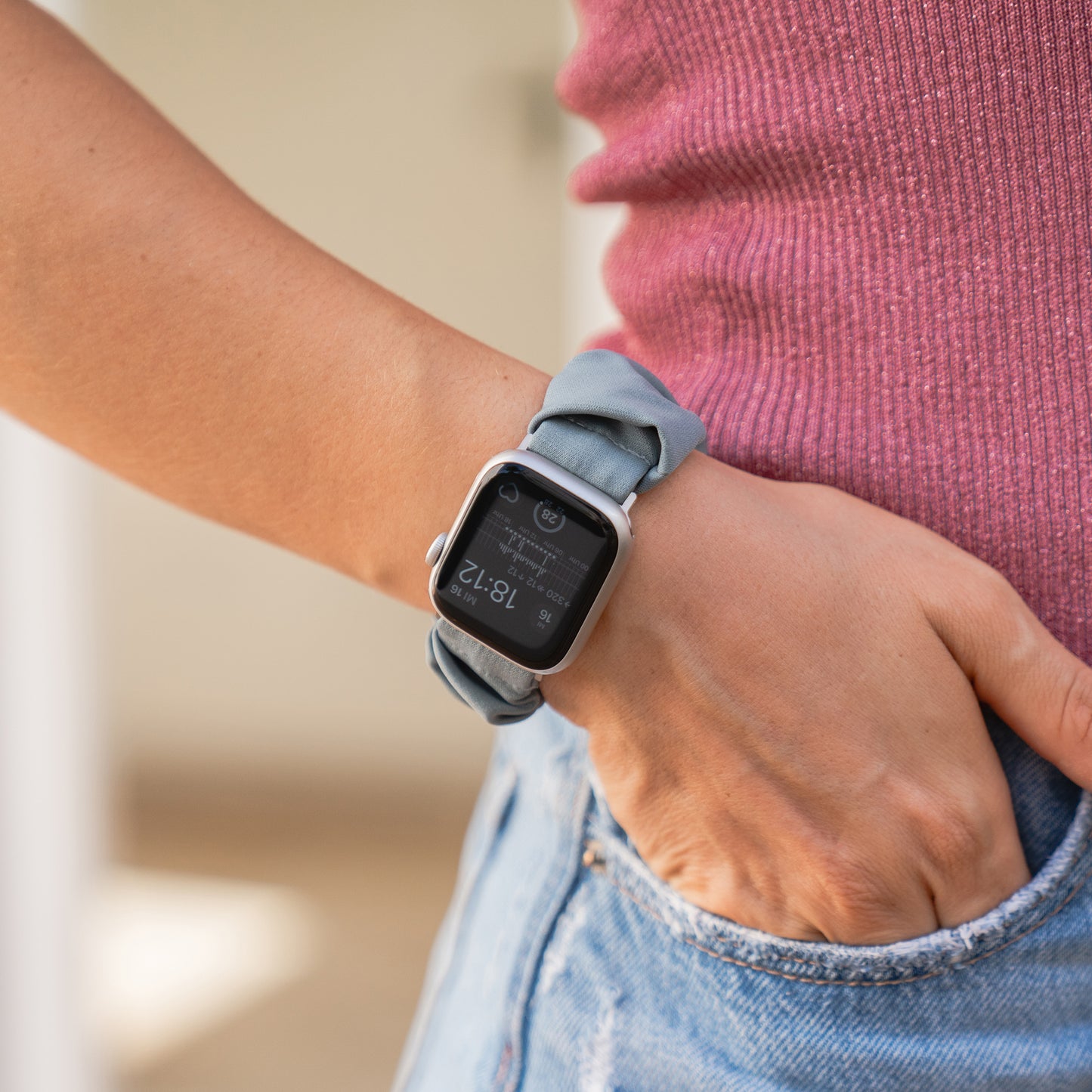 Eine Person in einem rosa Oberteil und Jeans trägt das arktisband Apple Watch Scrunchie Armband "Elanora" von arktisband mit einem grauen Band am linken Handgelenk.