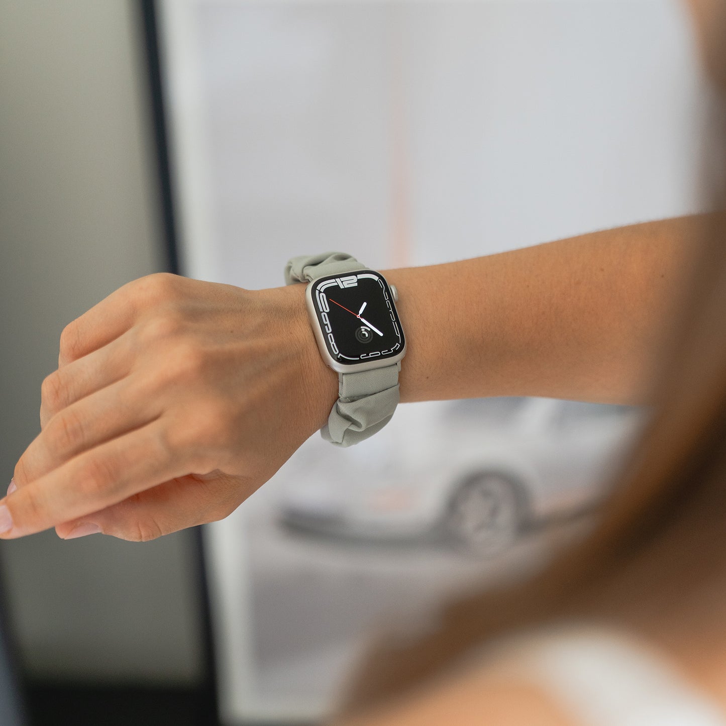 Eine Person, die ein arktisband Apple Watch Scrunchie Armband "Elanora" mit einem hellgrauen Band trägt, hält ihren Arm vor einem unscharfen Hintergrund aus.