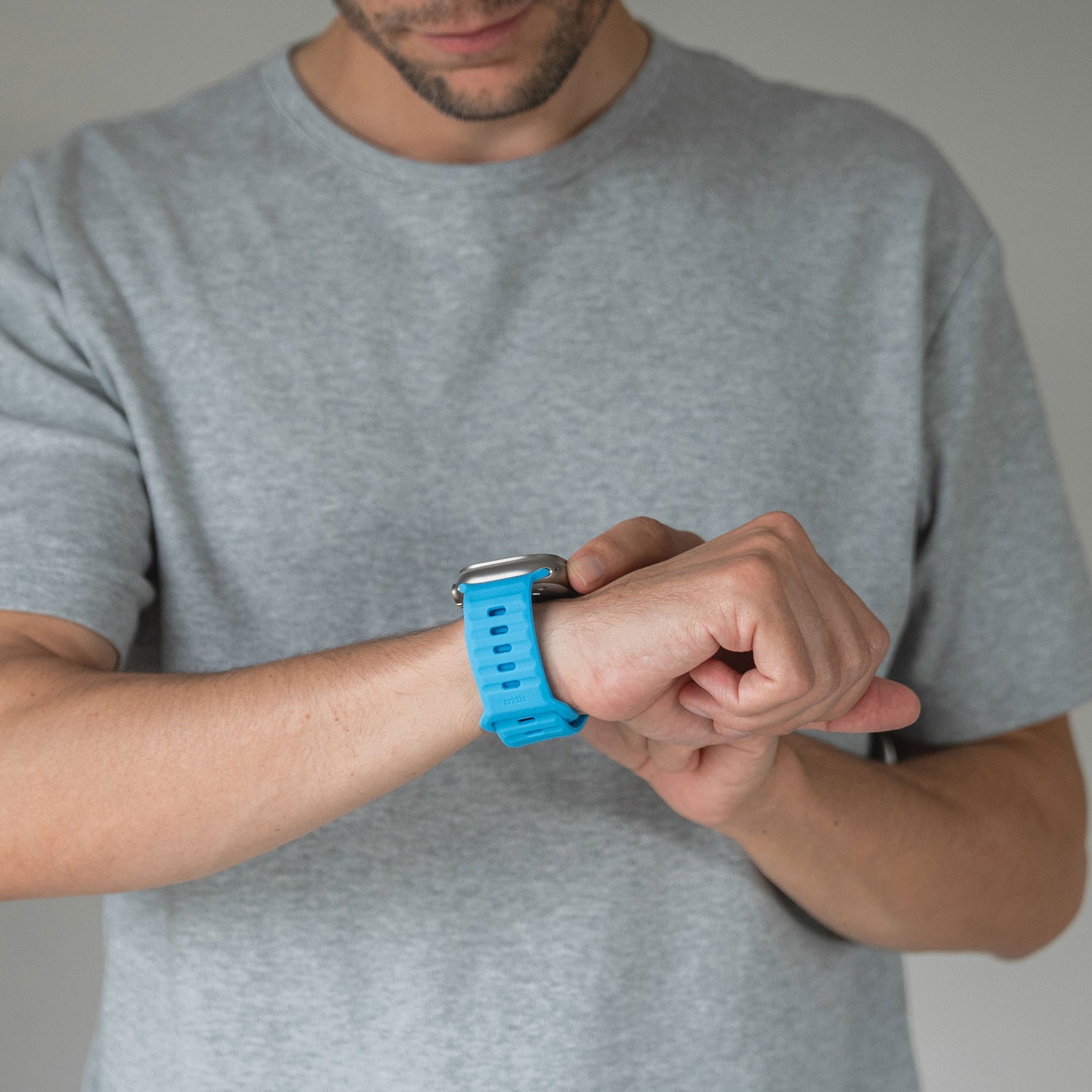 Eine Person in einem hellgrauen T-Shirt berührt mit der rechten Hand ihr Arktisband Apple Watch Silikonarmband „Active“ und genießt den angenehmen Tragekomfort und die Atmungsaktivität des blauen Arktisband-Armbands an ihrem linken Handgelenk.