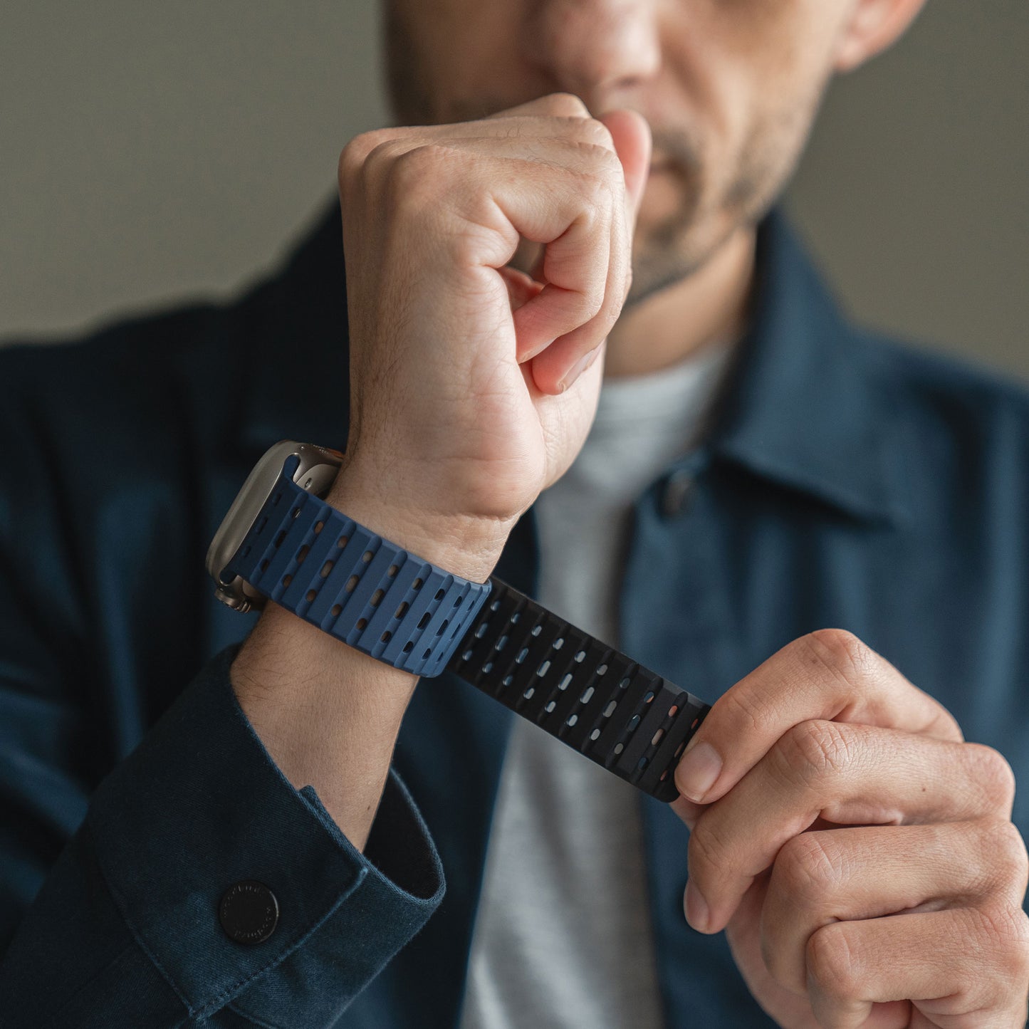Eine Person in grauem Hemd und dunkelblauer Jacke befestigt das magnetische Silikonarmband „Power“ (blau/schwarz) der Apple Watch, ihr Gesicht ist teilweise außerhalb des Bildes.