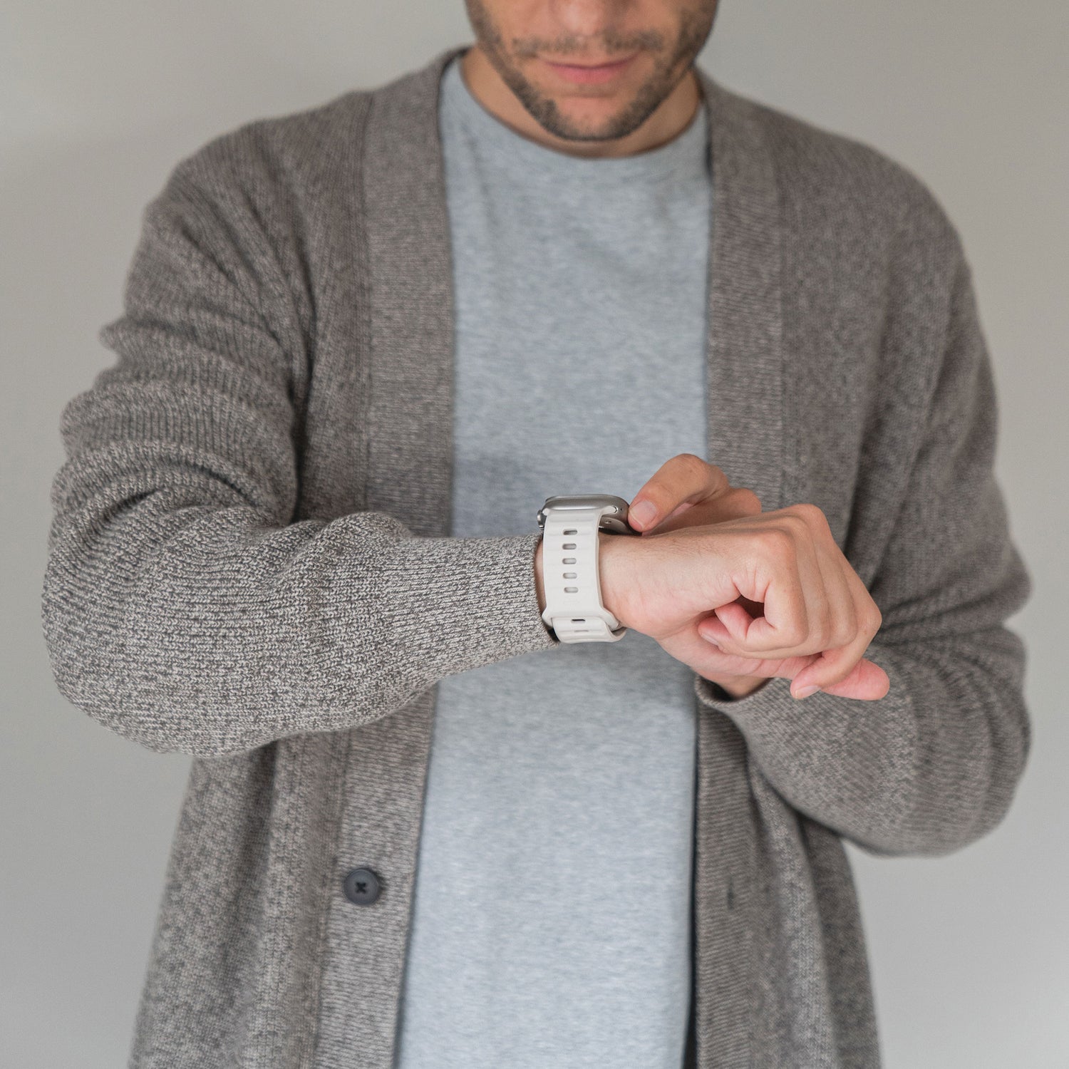 Jemand in einer grauen Strickjacke und einem hellgrauen Hemd trägt ein Arktisband Apple Watch Silikonarmband „Active“ am linken Handgelenk und steht vor einem schlichten, hellen Hintergrund.