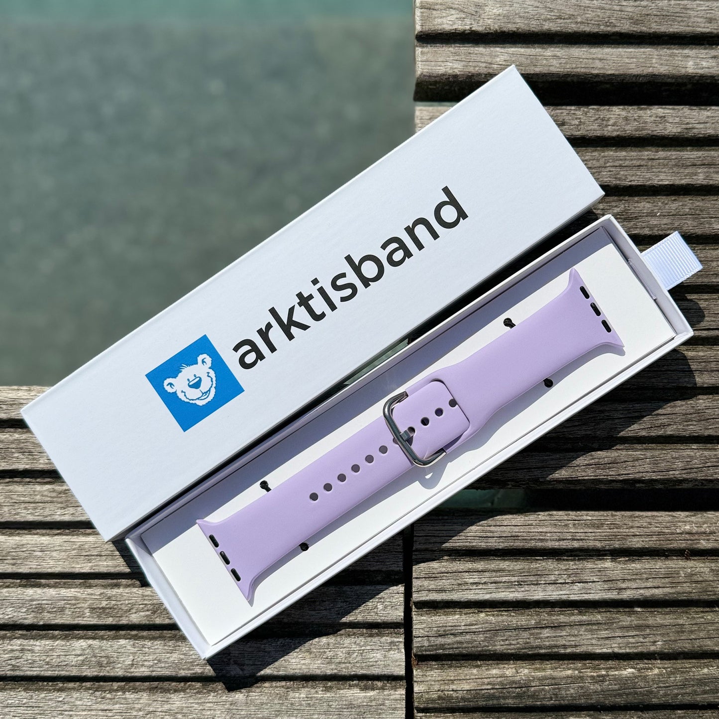 Das arktisband Apple Watch Silikonarmband "Aqua", ein hellviolettes Armband mit silberner Schließe, wird in einer offenen weißen Arktisband-Box auf Holz in der Nähe von Wasser präsentiert. Kompatibel mit der Apple Watch.