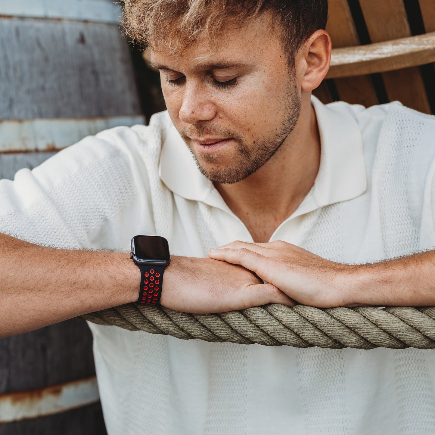 Ein hellbraunhaariger Mann mit kurzem Bart trägt ein weißes strukturiertes Poloshirt und das arktisband Apple Watch Silikonarmband „Energy“ von arktisband im schwarz-roten Design und ruht mit den Armen im Freien auf einem dicken Seil.