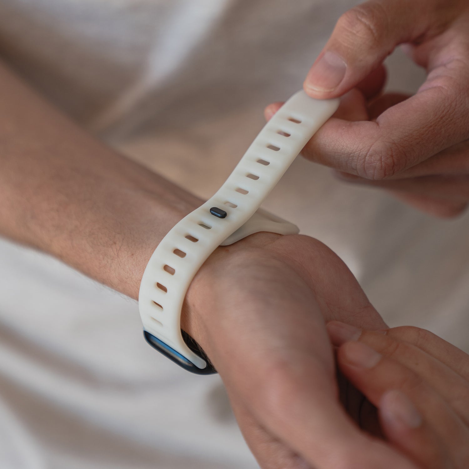 Eine Person befestigt mit beiden Händen ein weißes Arktisband Apple Watch Silikonarmband „Active“ mit atmungsaktivem Design am linken Handgelenk. Der Hintergrund ist unscharf.