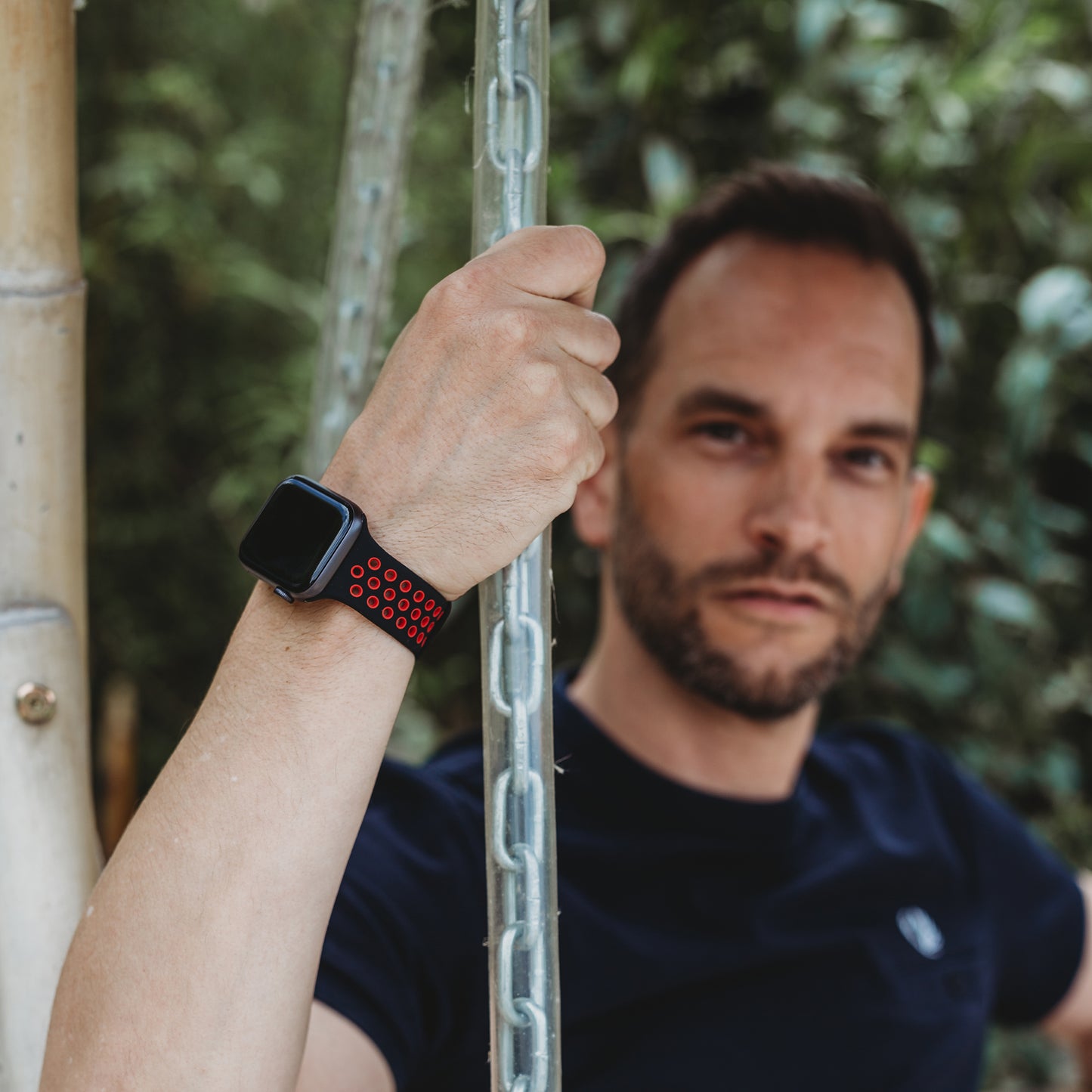 Ein Mann mit kurzen Haaren und Bart hält eine Metallkette in seiner linken Hand und trägt das arktisband Apple Watch Silikonarmband „Energy“ von arktisband. Das schwarz-rote perforierte Armband fällt auf, als er im Freien inmitten üppigen Grüns posiert.