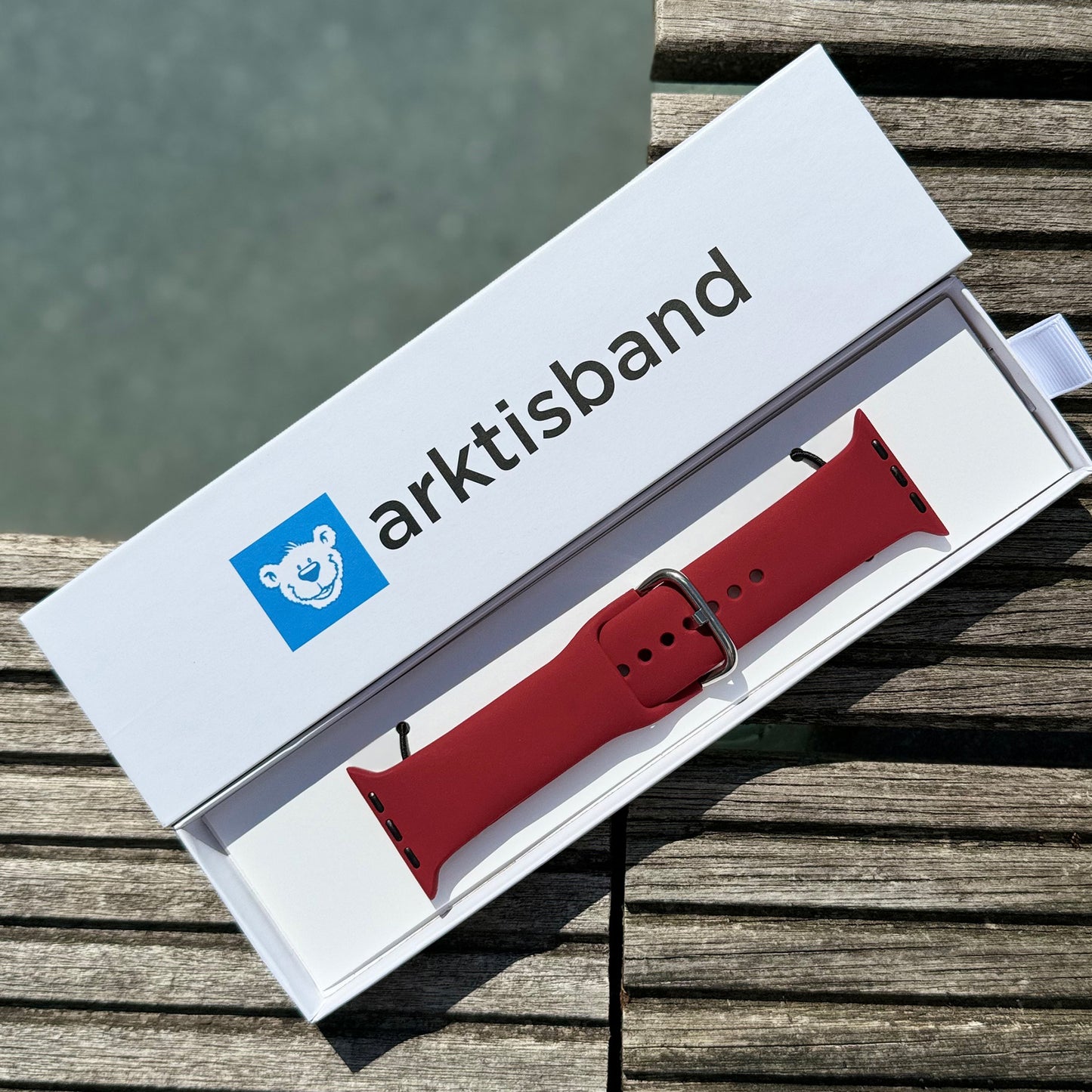 Das arktisband Apple Watch Silikonarmband „Aqua“ von arktisband liegt in einer weißen Box mit blauem Eisbär-Logo auf Holzlatten vor grauem, unscharfem Hintergrund. Das sportliche Armband ist Apple Watch kompatibel.
