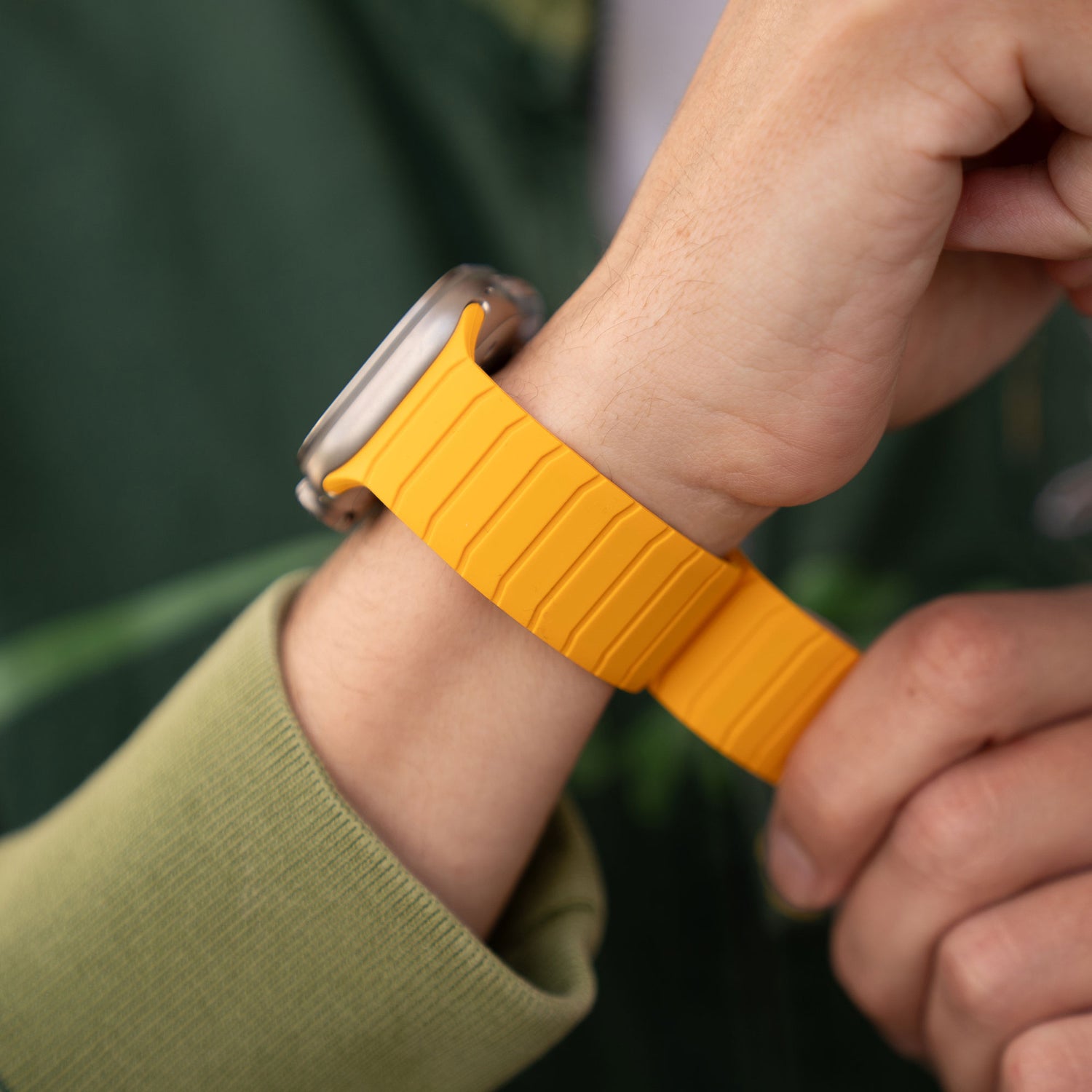 Eine Person passt ein magnetisches Armband „MagFlow“ für die Apple Watch mit gelbem, geschmeidigem Silikonband an ihrem Handgelenk an, während sie eine grüne Jacke und ein hellgrünes Sweatshirt trägt.