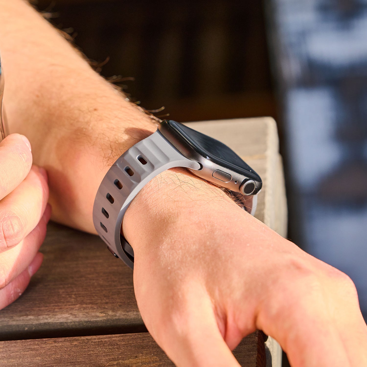 Jemand trägt ein arktisband Apple Watch Silikonarmband "Active" in Grau-Schwarz im sportlichen Look und legt seinen Arm auf Holz. Das Display ist leer, in der anderen Hand hält er einen Gegenstand.