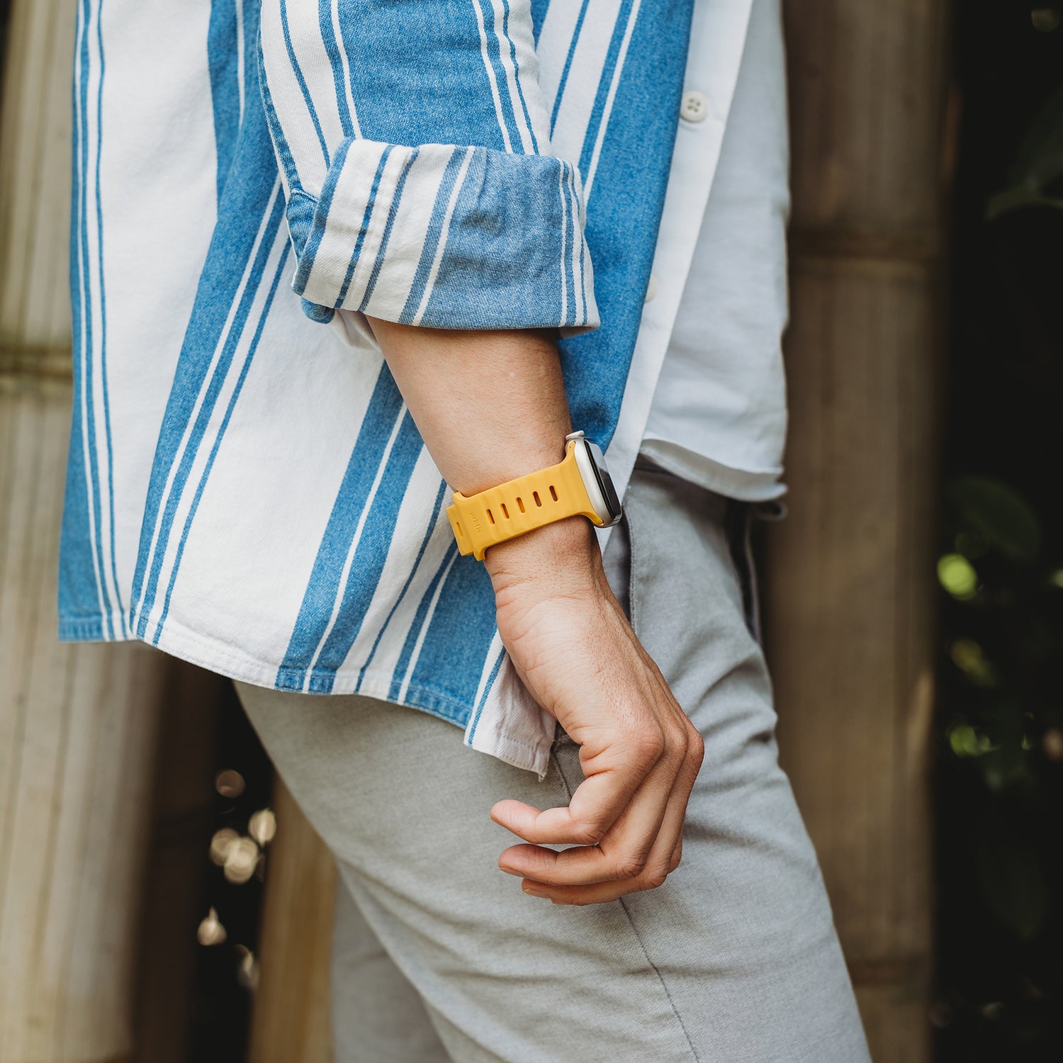Eine Person in einem blau-weiß gestreiften Hemd und einer hellgrauen Hose trägt das arktisband Apple Watch Silikonarmband "Active" von arktisband im atmungsaktiven Design. Im Hintergrund sind Bambusstäbe zu sehen.
