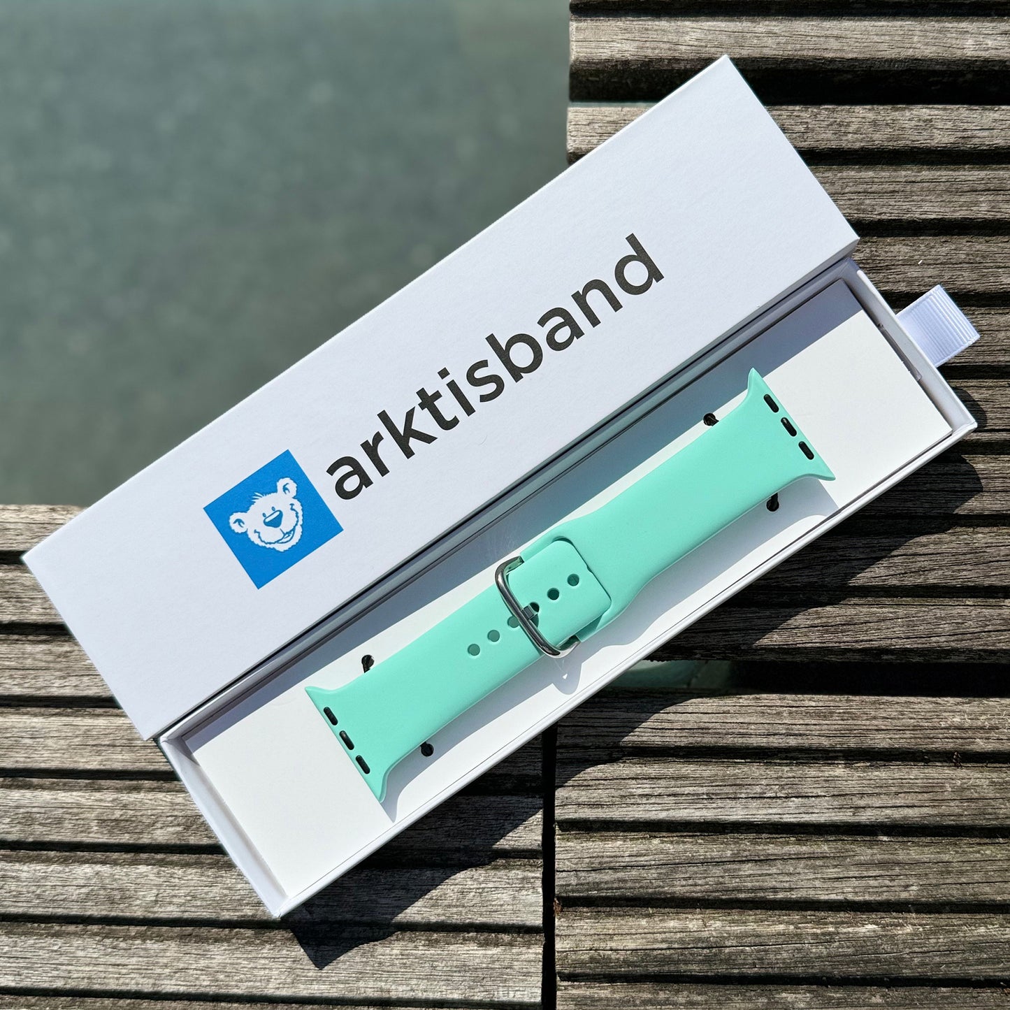 Das arktisband Apple Watch Silikonarmband „Aqua“, ein hellgrünes Sportarmband mit silberner Schnalle, wird in einer weißen arktisband-Box mit Eisbär-Logo gezeigt, die auf Holzlatten vor grauem Hintergrund ruht.