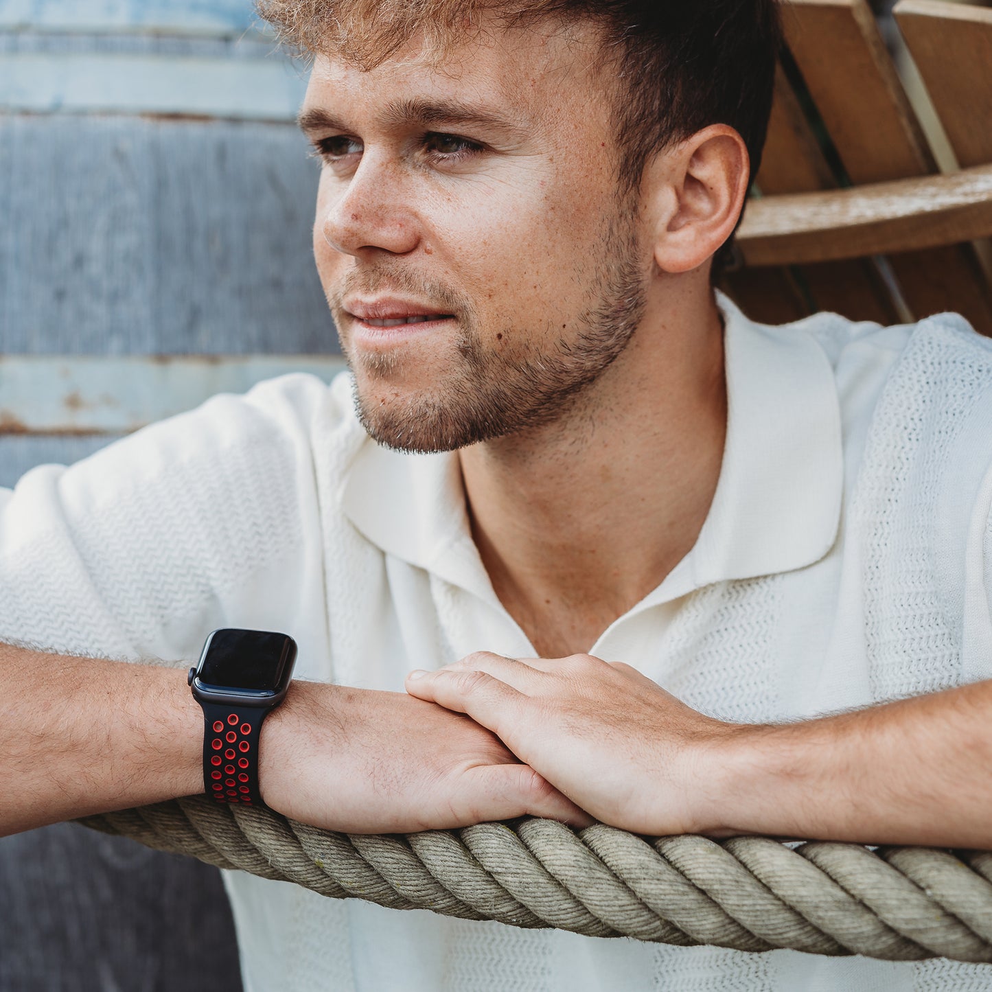Ein Mann mit kurzen Haaren und gestutztem Bart lehnt an einem dicken Seil. Er trägt ein weißes strukturiertes Kurzarmhemd und das Arktisband Apple Watch Silikonarmband "Energy" in Schwarz und Rot. Im Hintergrund sind Holzbretter zu sehen.