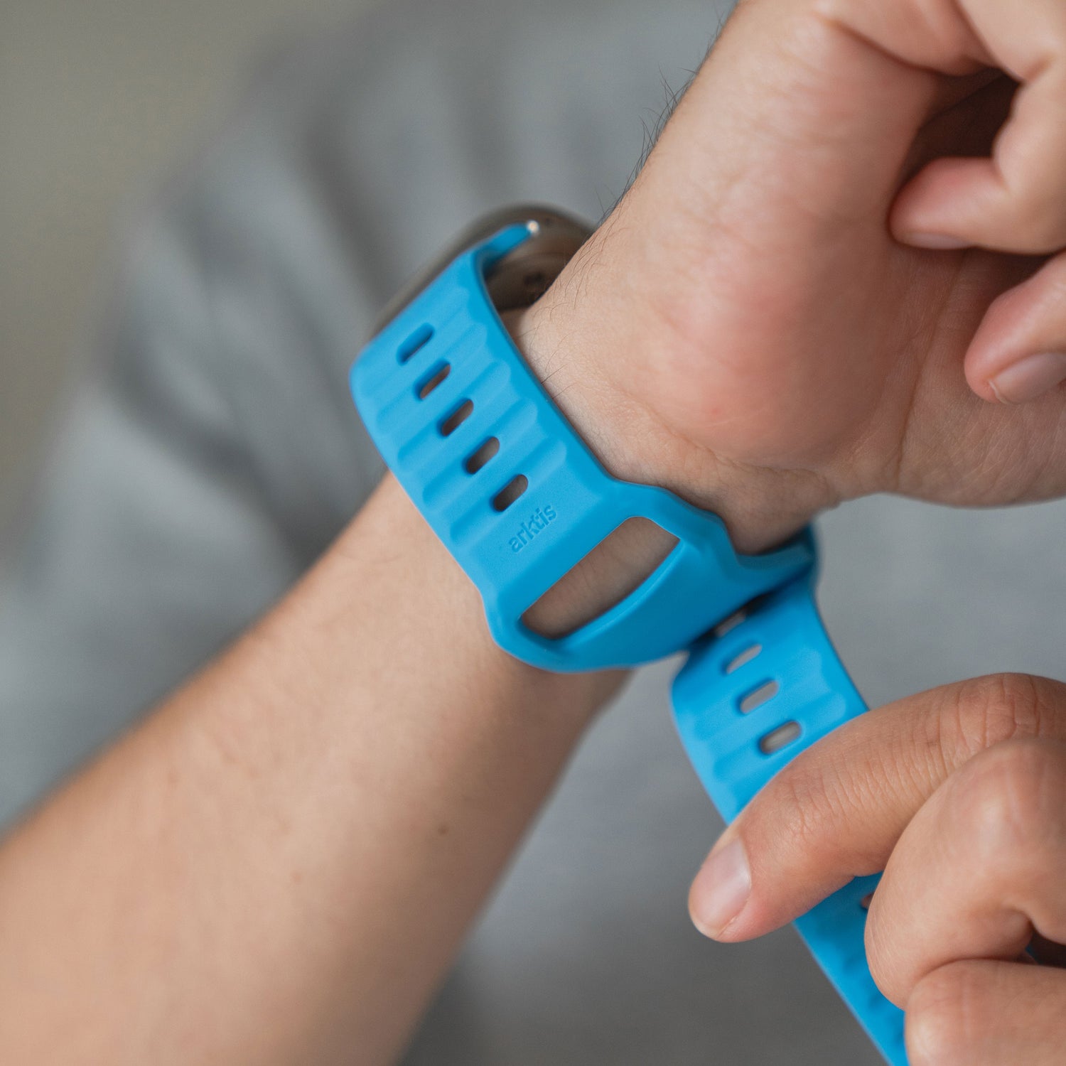 Eine Person befestigt das arktisband Apple Watch Silikonarmband "Active" in Blau mit rechteckigen Perforationen am linken Handgelenk und trägt ein graues Shirt. Das sportliche Armband von arktisband verleiht jeder Apple Watch einen modernen Look.