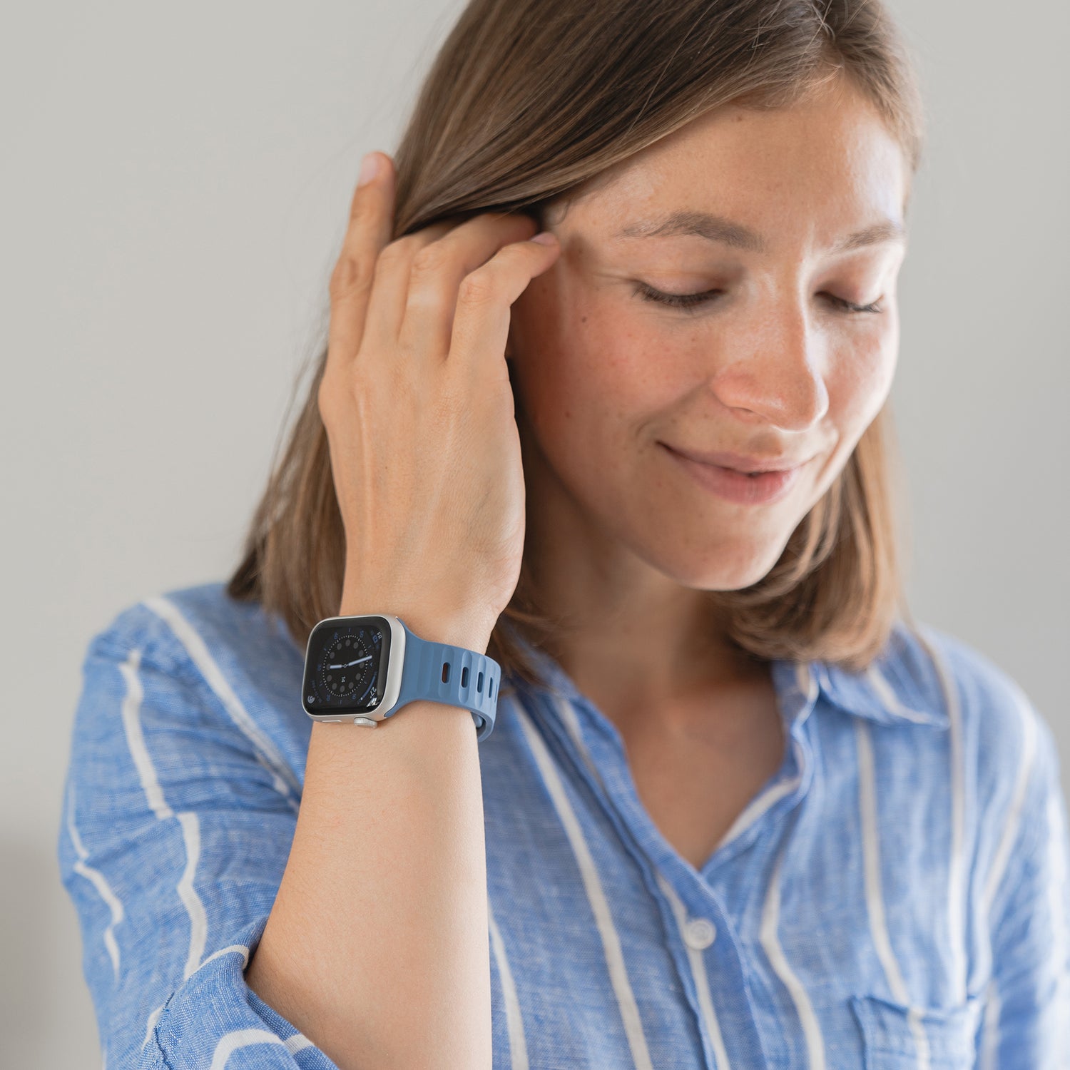 Eine Person, die ein blau-weiß gestreiftes Hemd und ein arktisband Apple Watch Silikonarmband „Active“ von arktisband trägt, berührt ihr Haar und blickt mit einem leichten Lächeln auf einen schlichten Hintergrund.