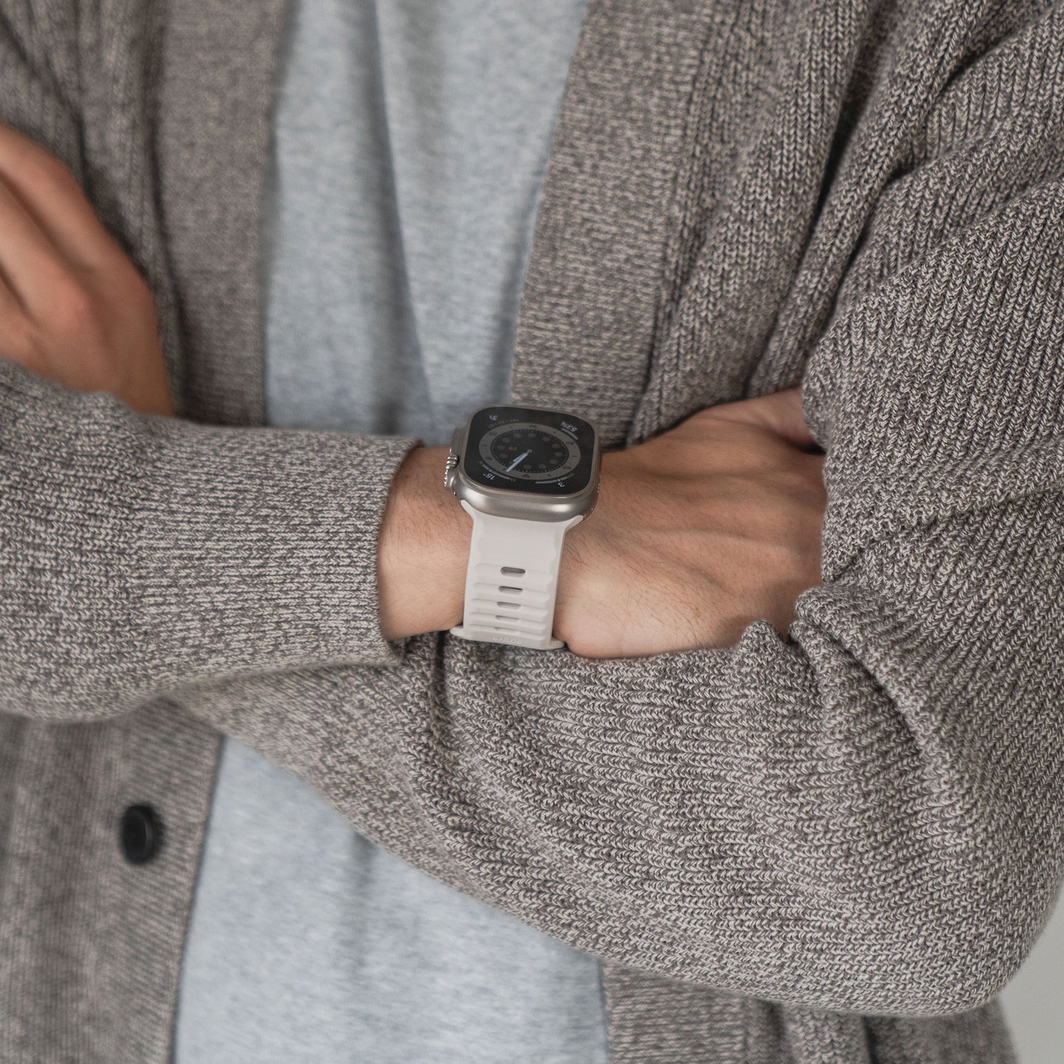 Eine Person mit verschränkten Armen trägt einen grauen Strickpullover und eine Apple Watch mit dem arktisband Apple Watch Silikonarmband "Active" von arktisband über einem hellgrauen Shirt. Nur Oberkörper und Arme sind sichtbar.
