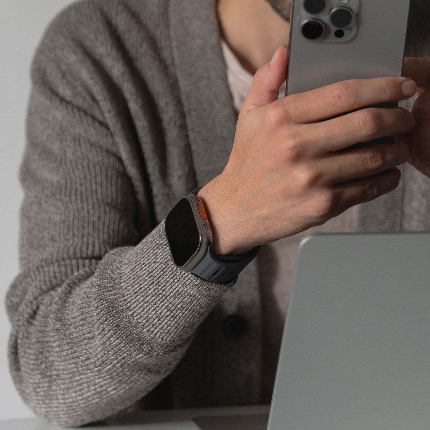 Eine Person im grauen Pullover hält ein Smartphone in der Hand und sitzt an einem Laptop. Ihr sportlicher Look wird durch ein arktisband Apple Watch Silikonarmband „Active“ von arktisband am Handgelenk komplettiert.