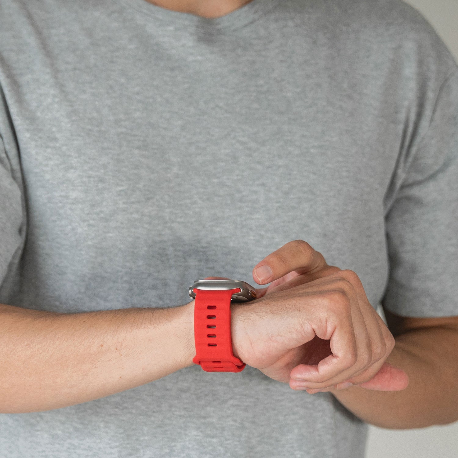 Eine Person in einem hellgrauen T-Shirt berührt vor einem schlichten, neutralen Hintergrund ein Arktisband Apple Watch Silikonarmband „Active“ mit rotem Band an ihrem linken Handgelenk.