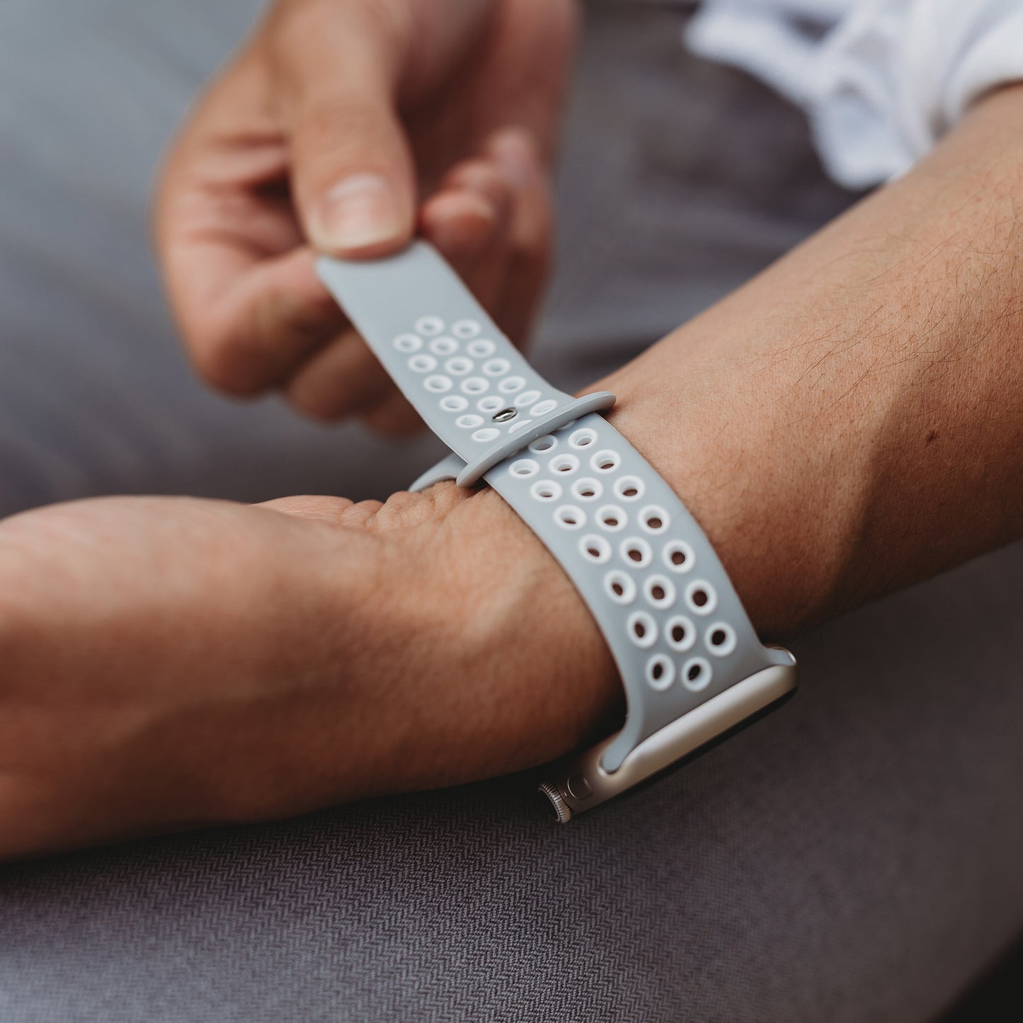 Eine Person befestigt ein Arktisband Apple Watch Silikonarmband "Energy" in Grau mit weißen Perforationen am Handgelenk. Hand und Unterarm sind scharf dargestellt und zeigen das Arktisband, während der Hintergrund unscharf ist.