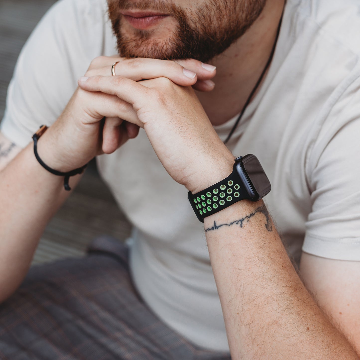 Eine bärtige Person im weißen Hemd stützt ihr Kinn auf die Hände und trägt ein arktisband Apple Watch Silikonarmband „Energy“ in Schwarz und Grün. An Unterarmen und Fingern sind Tattoos und ein Ring zu sehen.