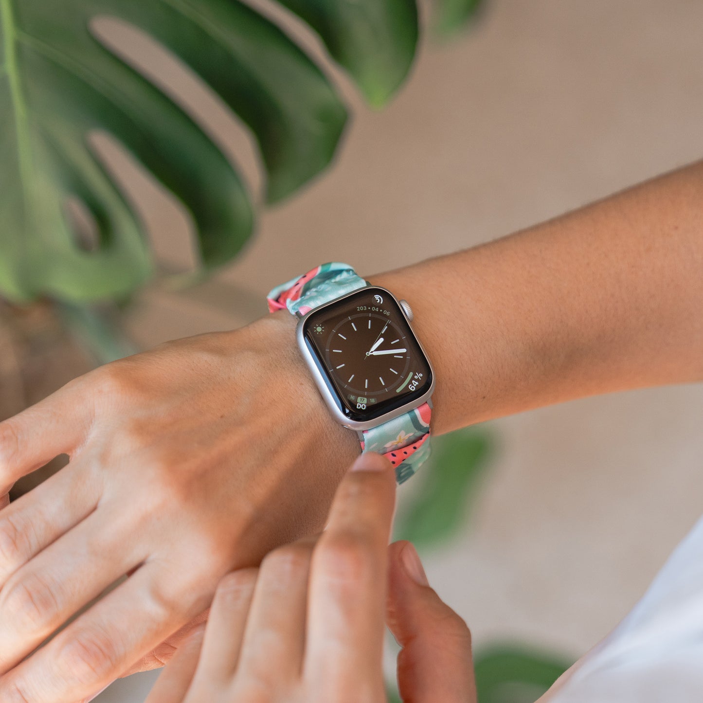 Jemand trägt das Arktisband Apple Watch Scrunchie Armband "Elanora", während er den Bildschirm berührt, mit einem grünen Blatt im Hintergrund.