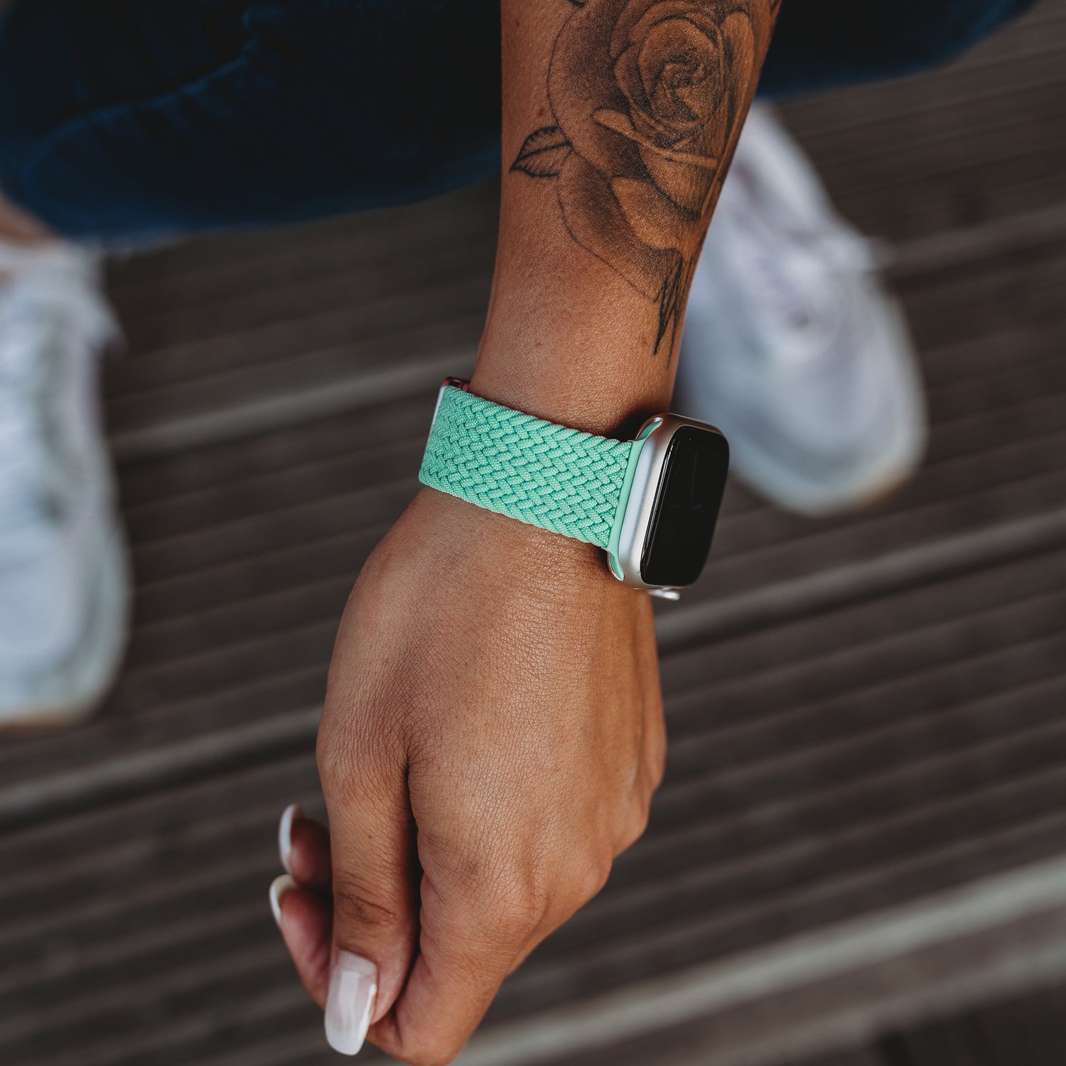 Eine Hand mit einem Rosentattoo trägt ein Arktisband Geflochtenes Apple Watch Armband „Casual“ mit Magnetverschluss in Mintgrün und steht auf Holzbrettern.