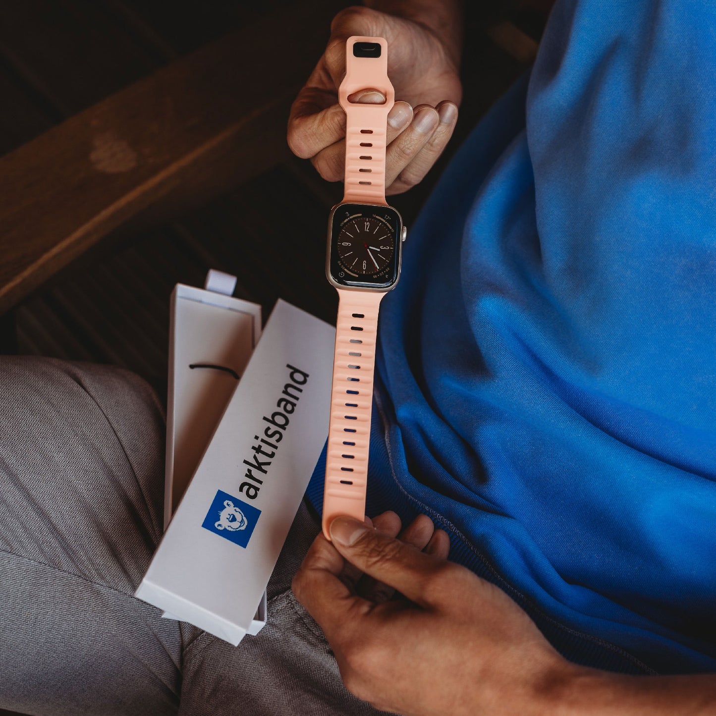 Eine Person im blauen Hemd hält das arktisband Apple Watch FKM Sportarmband „Sweat“ mit pfirsichfarbenem Armband. Die weiße, geöffnete Schachtel mit der Aufschrift „arktisband, Apple Watch kompatibel“ liegt auf ihrem Schoß, das Armband ist teilweise sichtbar.