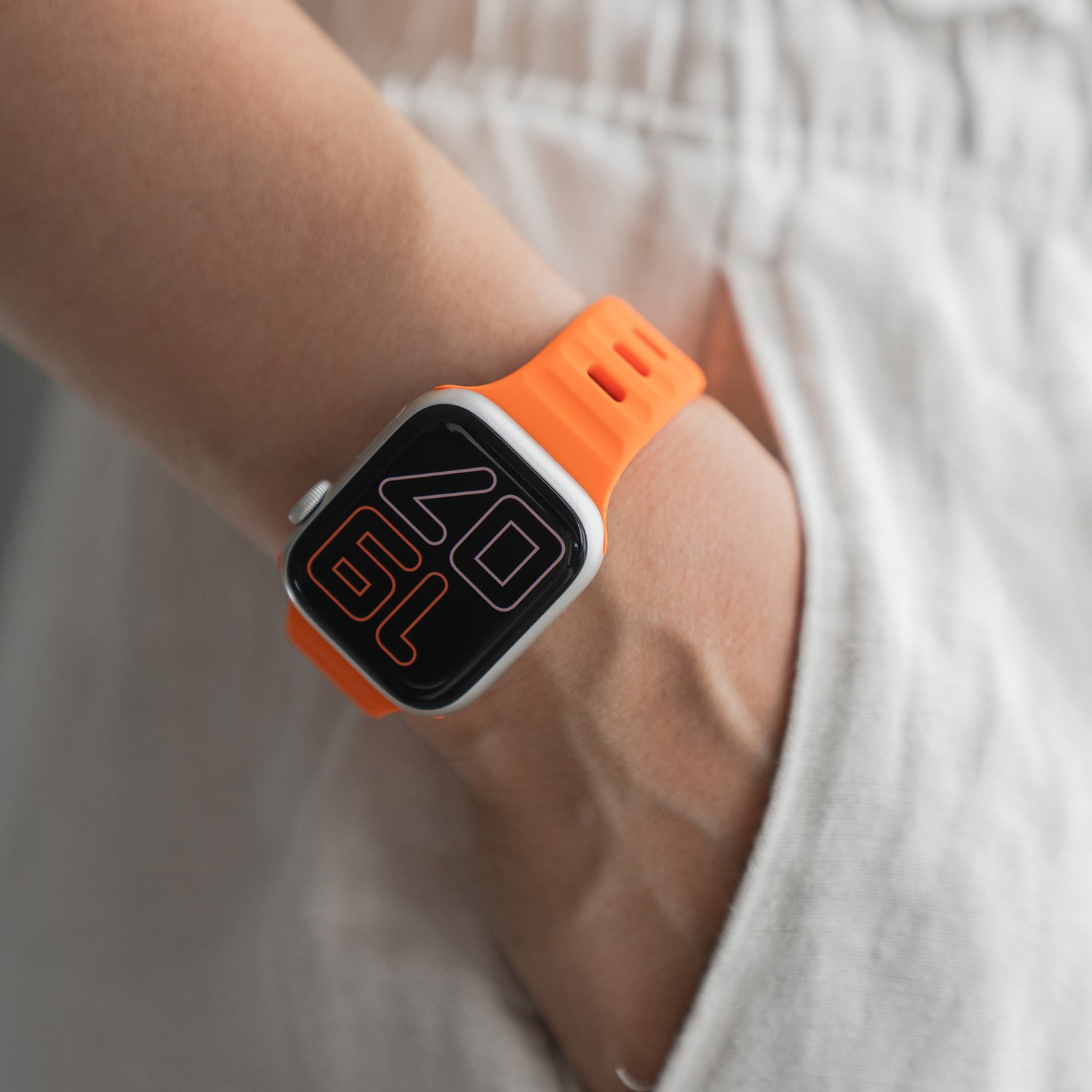 Eine Person in weißer Hose hat die Hand in der Tasche und präsentiert das arktisband Apple Watch Silikonarmband „Active“ von arktisband mit orangefarbenem Band und digitaler Anzeige.