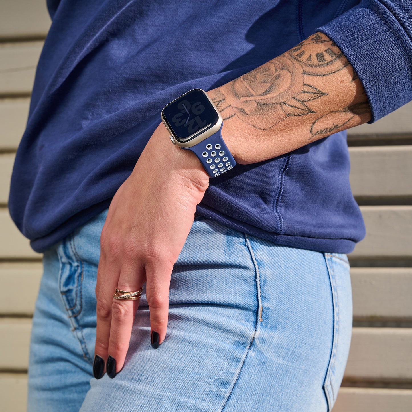 Eine Person in hellblauer Jeans und einem marineblauen Sweatshirt steht mit einer Hand auf der Hüfte und trägt das arktisband Apple Watch Silikonarmband "Energy" von arktisband. Ihr Arm zeigt ein Blumentattoo und sie hat schwarzen Nagellack.