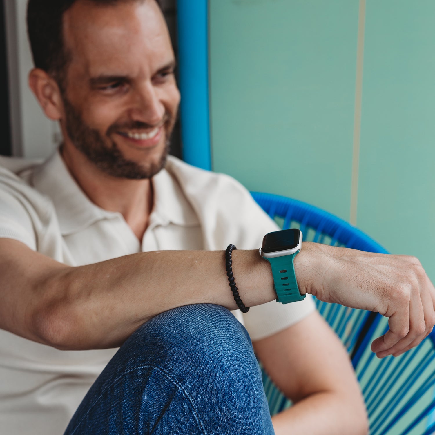 Ein Mann in weißem Poloshirt und Blue Jeans sitzt lächelnd auf einem blauen Stuhl und präsentiert das arktisband Apple Watch Silikonarmband „Active“ mit atmungsaktivem Design von arktisband. Außerdem trägt er ein schwarzes Perlenarmband.