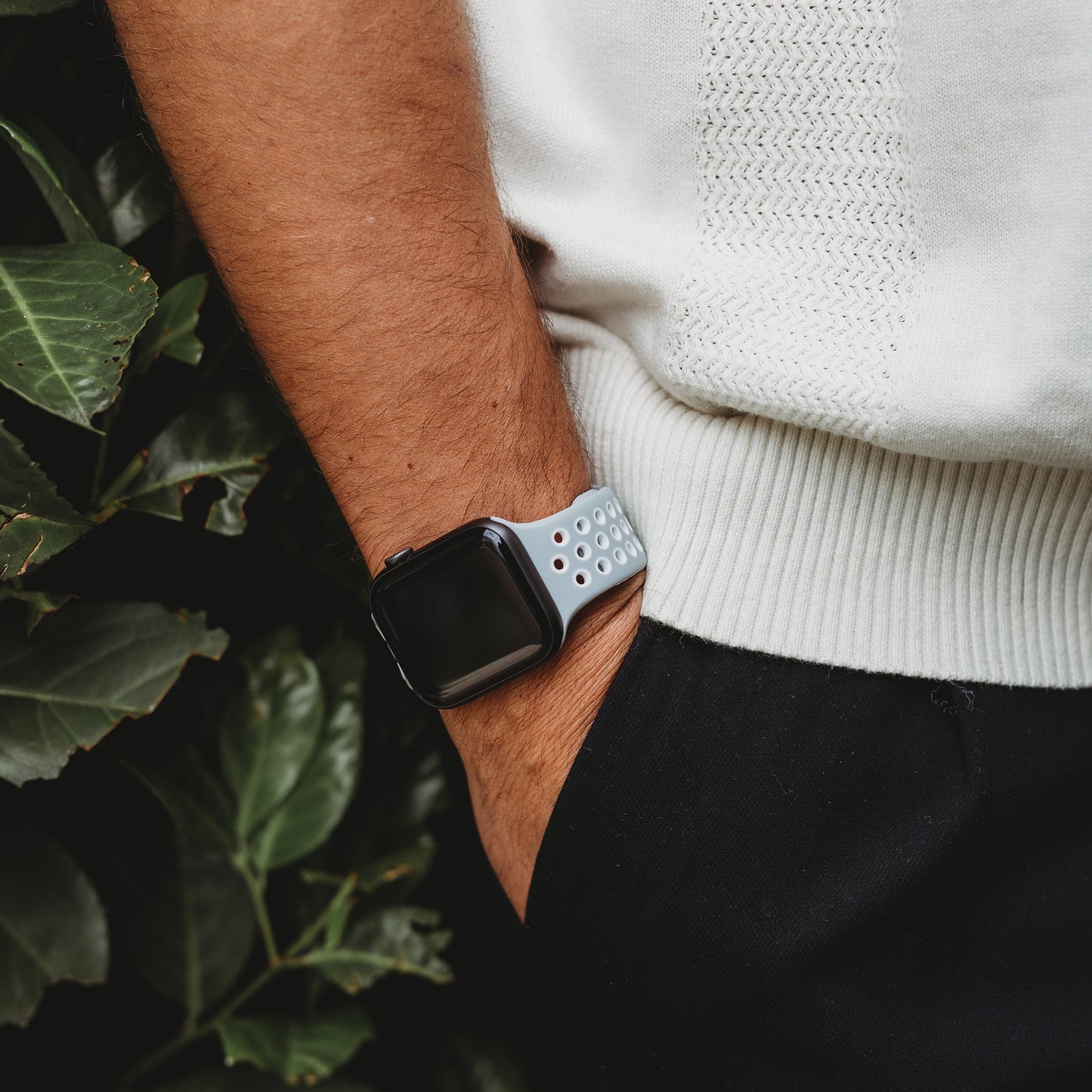 Eine Person, die ein weißes strukturiertes Hemd, eine schwarze Hose und das Arktisband Apple Watch Silikonarmband „Energy“ trägt, steht mit der Hand in der Tasche neben grünen Blattpflanzen.