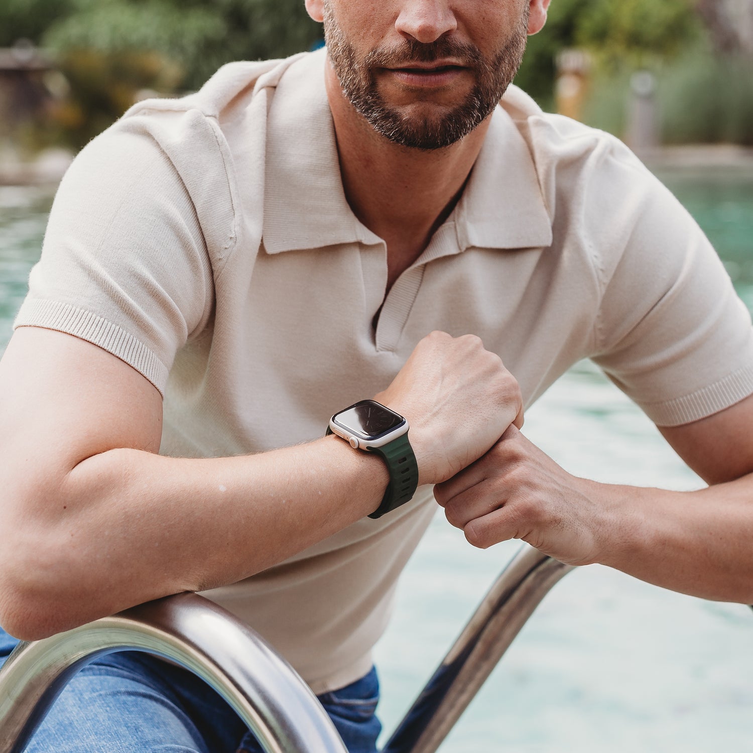 Ein Mann in einem beigen Kurzarm-Poloshirt und einem Arktisband Apple Watch Silikonarmband „Active“ sitzt am Pool und hält sich mit einem Arm an einer Metallleiter fest.