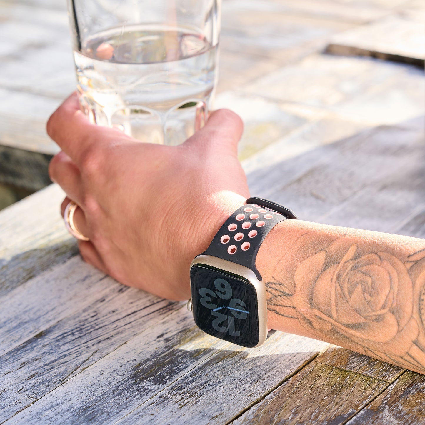 Eine Person mit floralem Unterarmtattoo und Ring hält ein Glas Wasser. An ihrem Handgelenk trägt sie ein arktisband Apple Watch Silikonarmband „Energy“ mit schwarz-pinkem Lochmuster der Marke arktisband.