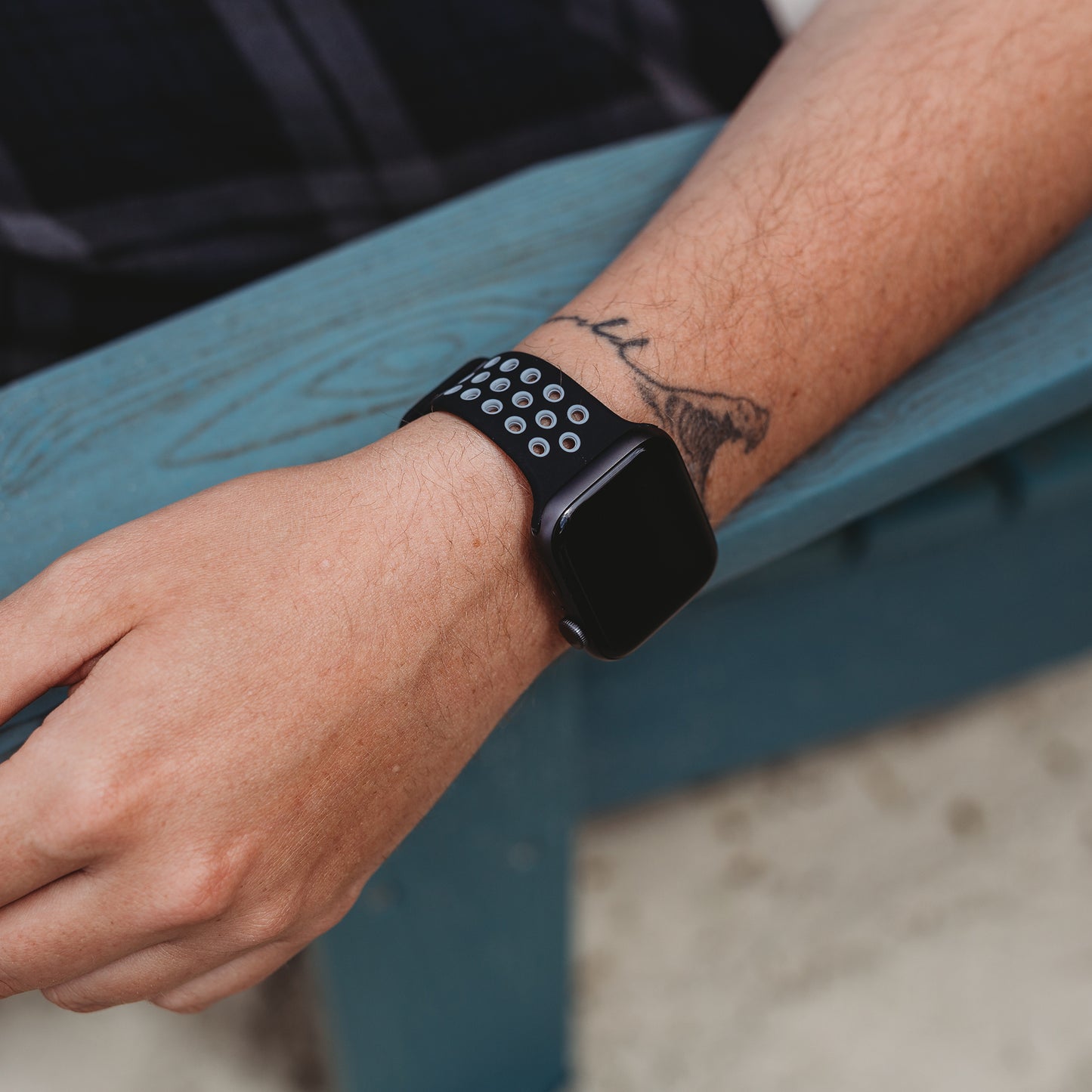 Eine Person legt ihren Arm auf eine blaue Fläche und trägt ein arktisband Apple Watch Silikonarmband „Energy“ von arktisband. Ein schwarzes Zweig-Tattoo ziert ihren Unterarm in der Nähe des perforierten Armbands der Smartwatch.