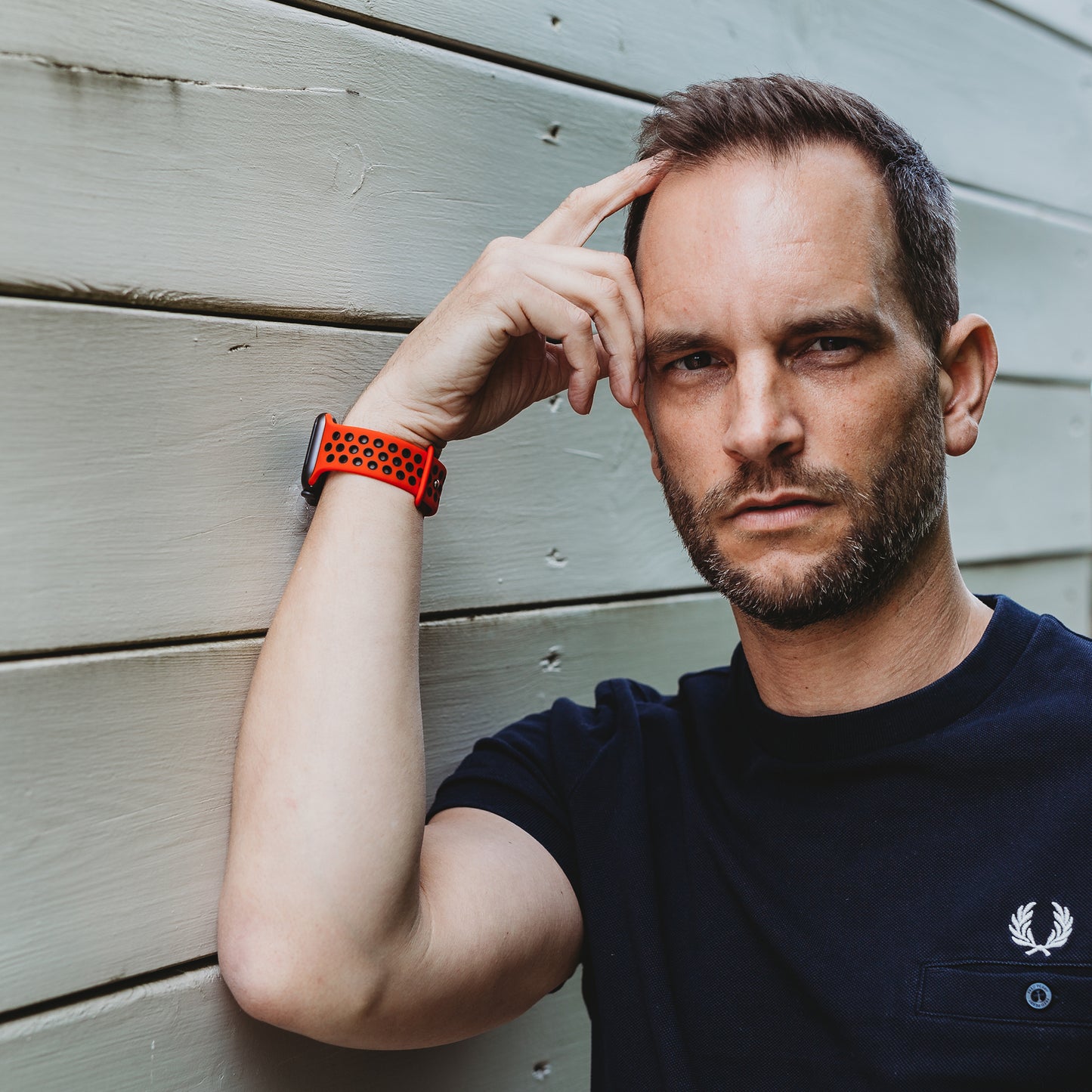 Ein Mann mit kurzen Haaren und Bart trägt ein schwarzes T-Shirt und das rote Arktisband Apple Watch Silikonarmband „Energy“, steht mit der Hand an der Schläfe vor einer hellen Holzwand und blickt in die Kamera.