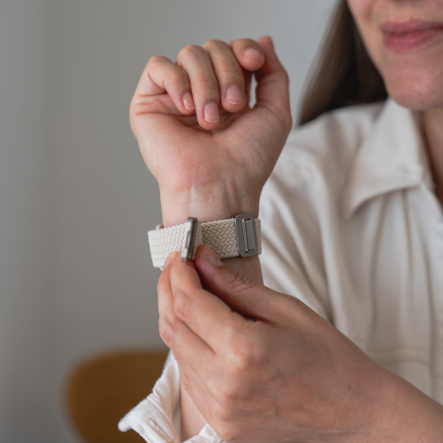 Eine Person passt das Arktisband Geflochtenes Apple Watch Armband "Casual" mit Magnetverschluss in Beige aus Polyestergarn an ihrem linken Handgelenk an und trägt dabei ein helles Langarmshirt. Nur die untere Gesichtshälfte ist sichtbar.