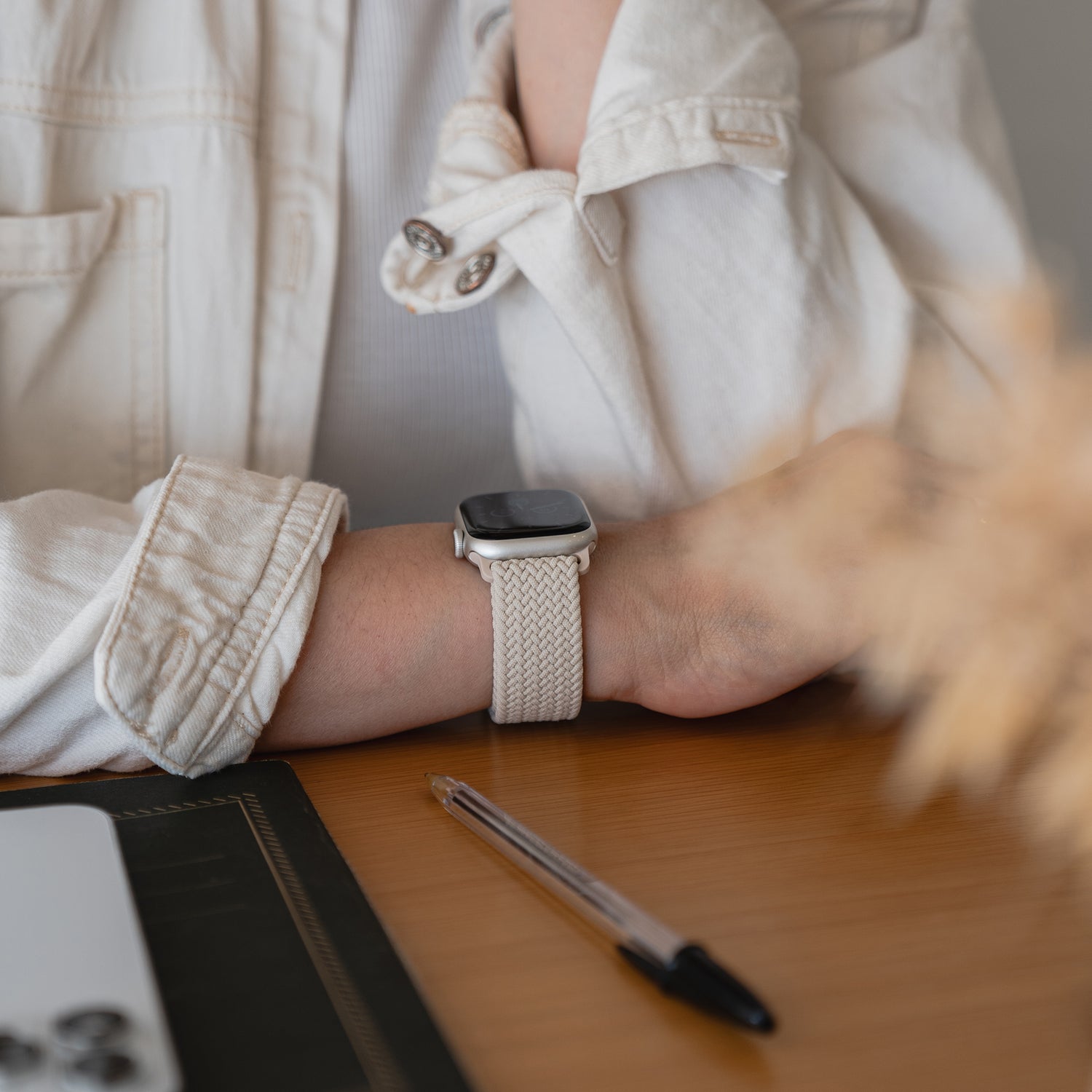 Eine Person, die eine cremefarbene Jacke und ein geflochtenes Apple Watch Armband „Casual“ mit Magnetverschluss trägt, legt ihren Arm neben einem Stift und einem Teil eines Laptops auf einen Holztisch.