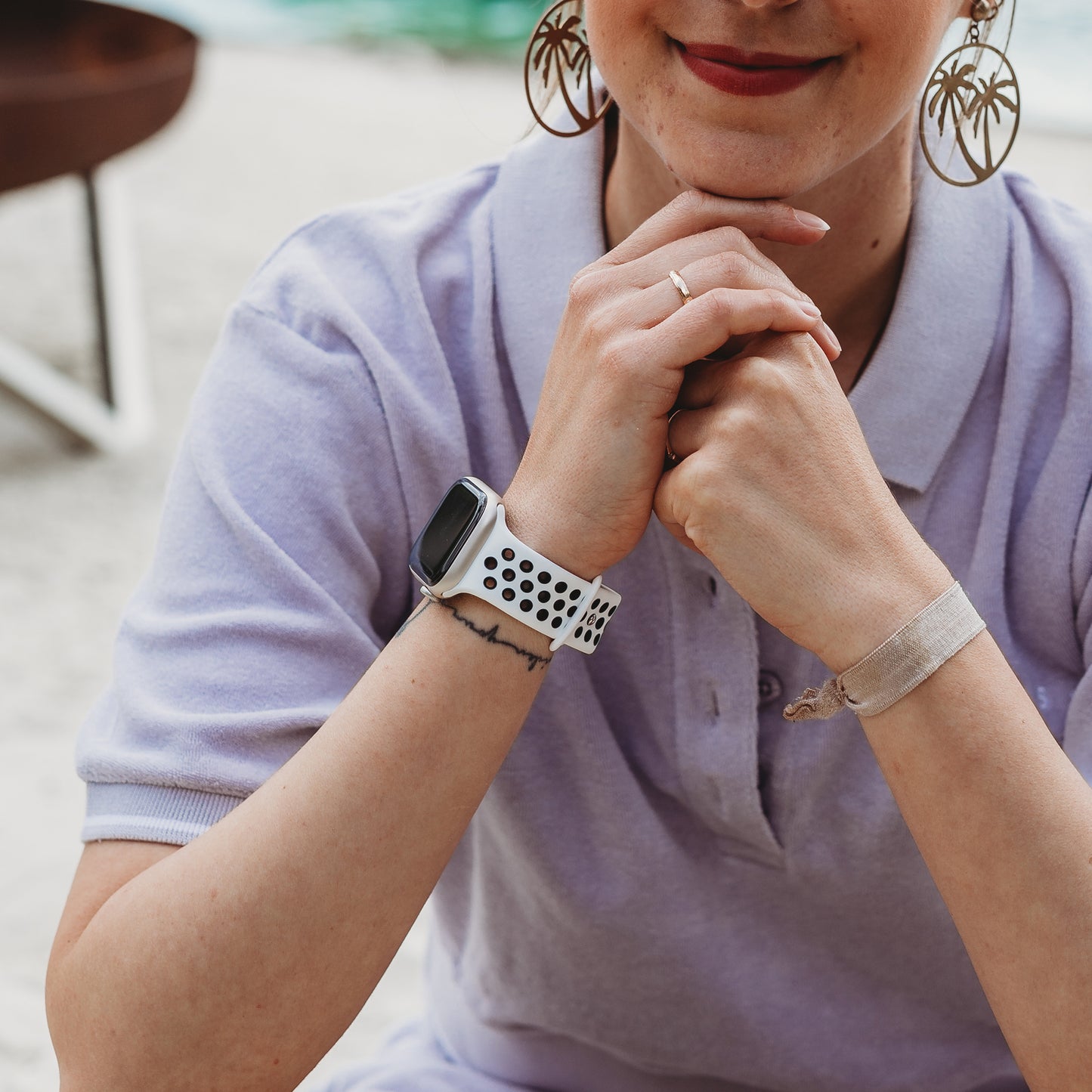 Eine Person in einem hellvioletten Poloshirt, Creolen und einem zarten Ring stützt ihr Kinn im Freien auf ihre Hände. Ihre Apple Watch ist mit dem eleganten arktisband Apple Watch Silikonarmband „Energy“ von arktisband vor einem sanft verschwommenen Hintergrund gestylt.