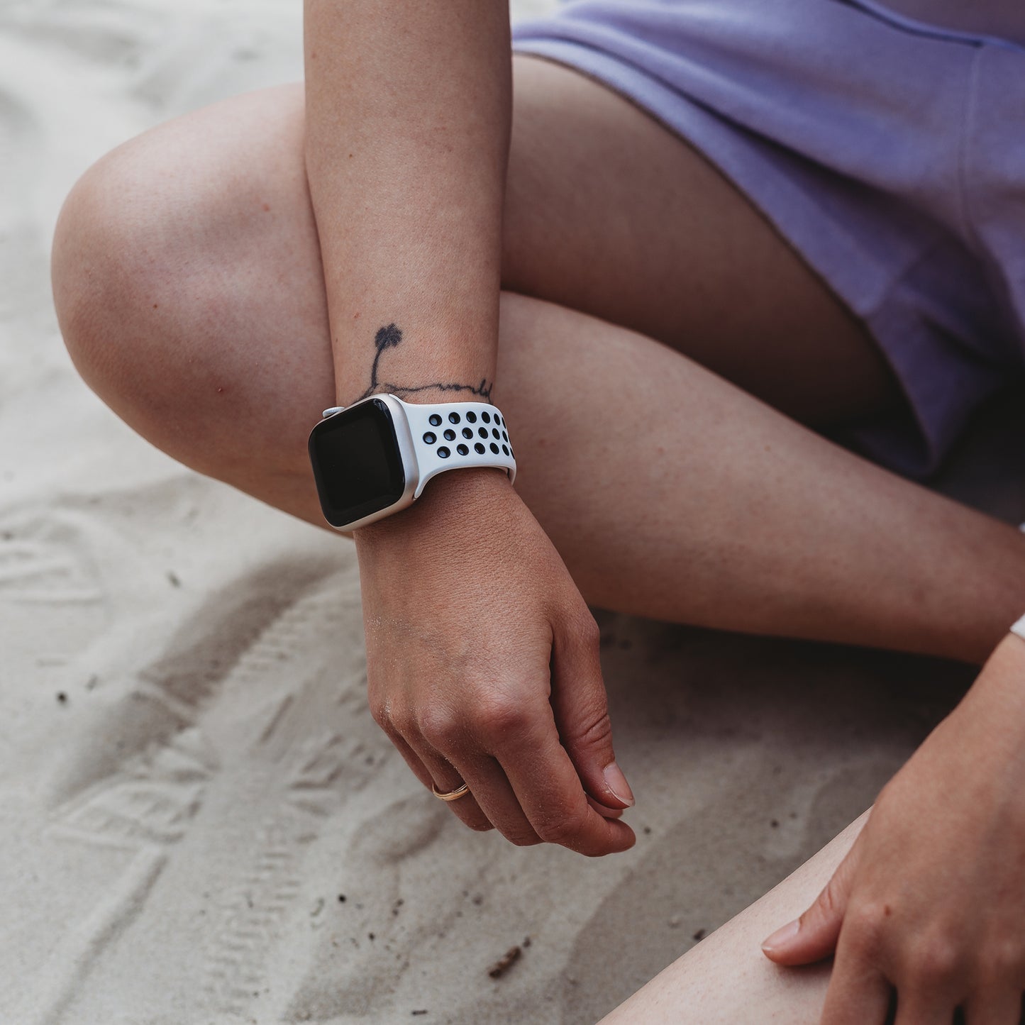 Jemand trägt das arktisband Apple Watch Silikonarmband „Energy“ von arktisband mit einem weiß-schwarzen perforierten Band und sitzt im Schneidersitz in hellvioletten Shorts auf dem Sand, wobei ein kleines Tattoo am Handgelenk sichtbar ist.