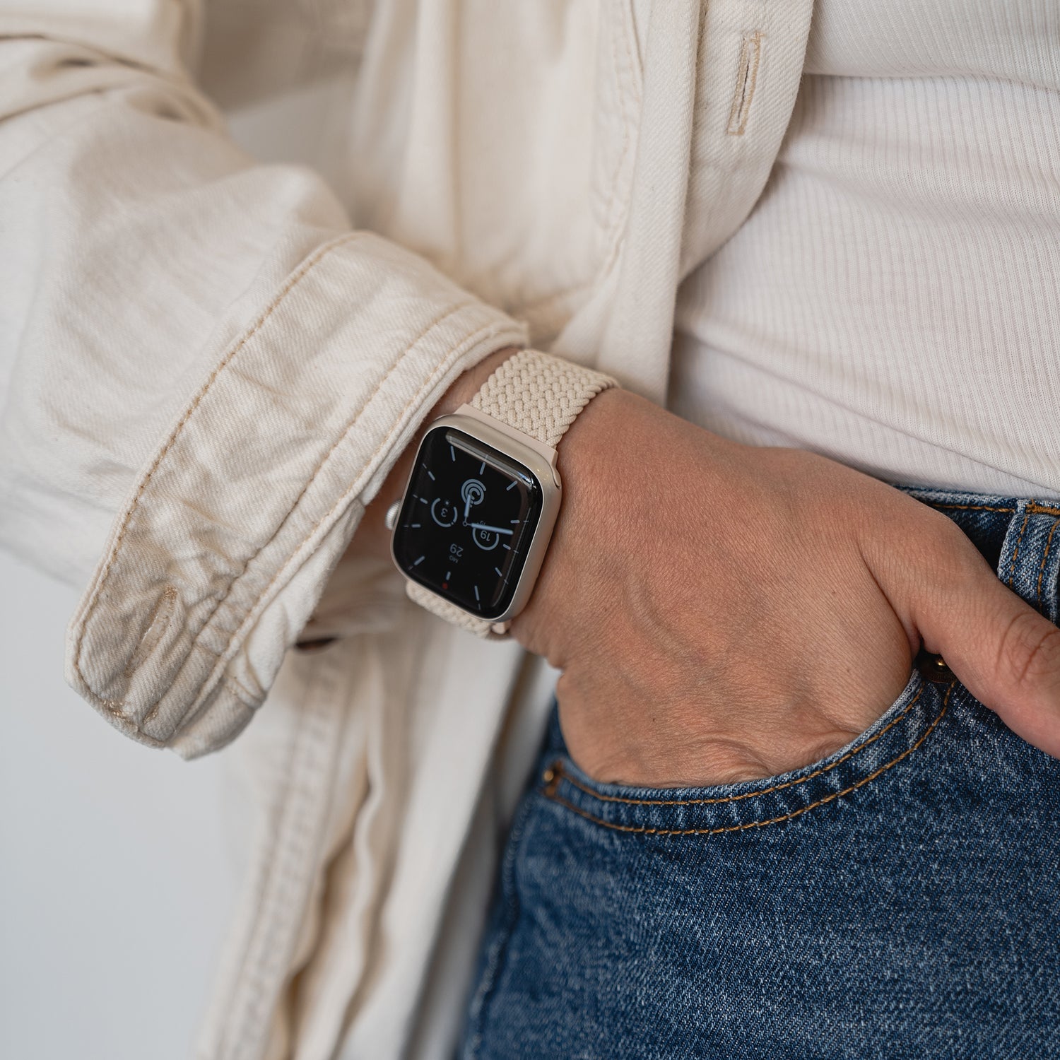 Eine Person trägt ein arktisband Geflochtenes Apple Watch Armband „Casual“ mit Magnetverschluss in Beige, dazu ein cremefarbenes Hemd und blaue Jeans. Die Hand steckt in der Tasche, und das Zifferblatt zeigt digitale Informationen.