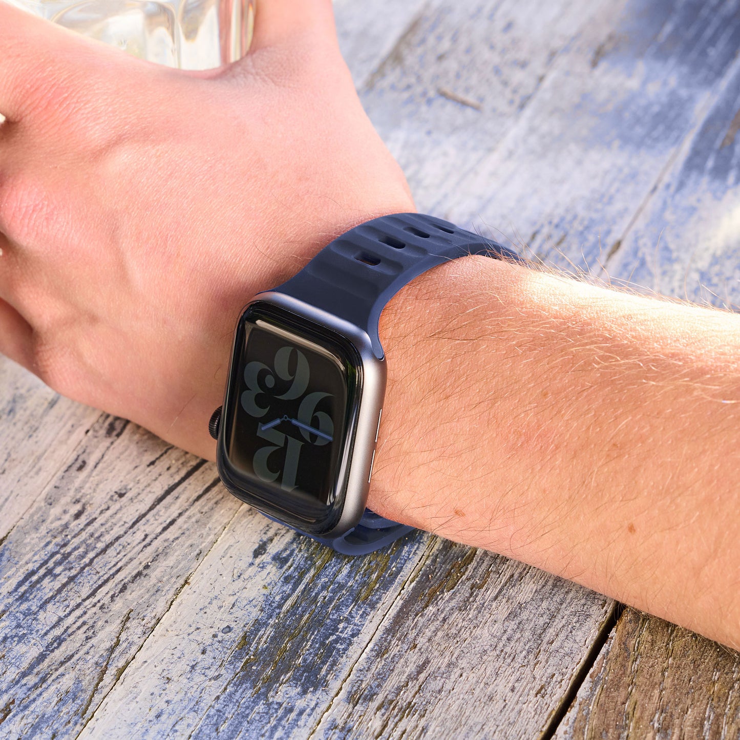 Jemand trägt ein arktisband Apple Watch Silikonarmband „Active“ von arktisband in Dunkelblau und hält ein Glasobjekt. Der sportliche Look wird durch den verwitterten Holzhintergrund und die große digitale Uhranzeige der Uhr unterstrichen.