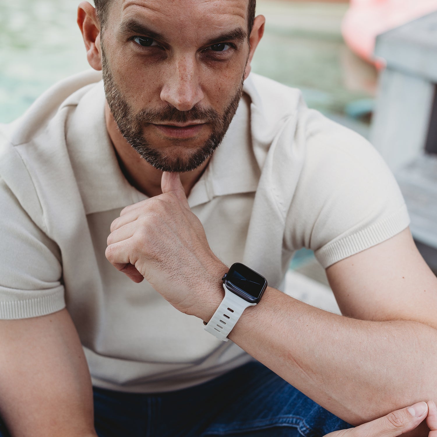 Ein Mann mit kurzen Haaren und Bart sitzt im Freien, trägt ein helles Kurzarmhemd und das arktisband Apple Watch Silikonarmband „Active“ mit atmungsaktivem Design am linken Handgelenk.