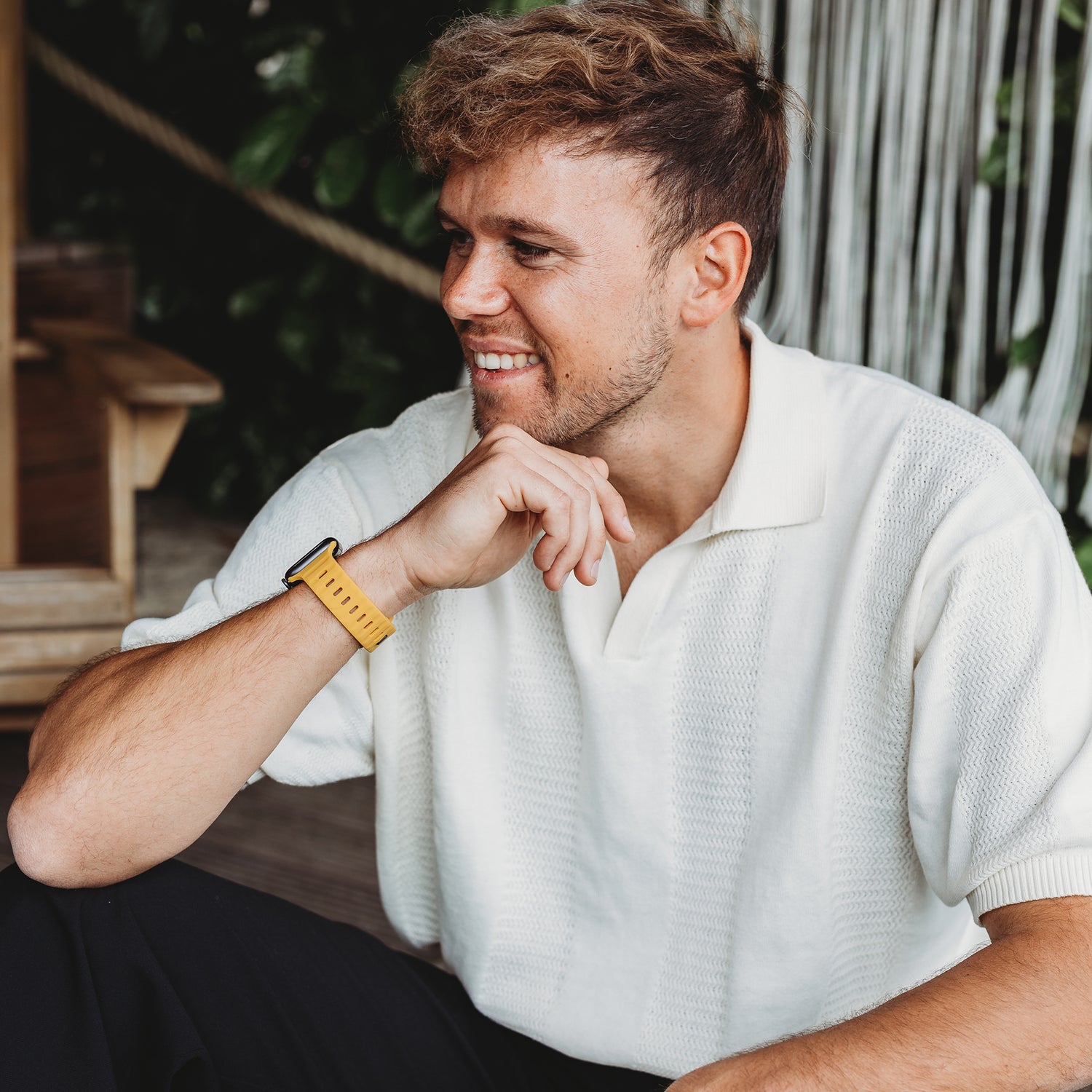Ein Mann mit kurzen, hellbraunen Haaren und gestutztem Bart sitzt da, die Hand am Kinn, lächelt und blickt zur Seite. Er trägt ein weißes strukturiertes Hemd und eine Apple Watch Ultra mit dem arktisband „Active“ Silikonarmband von arktisband.