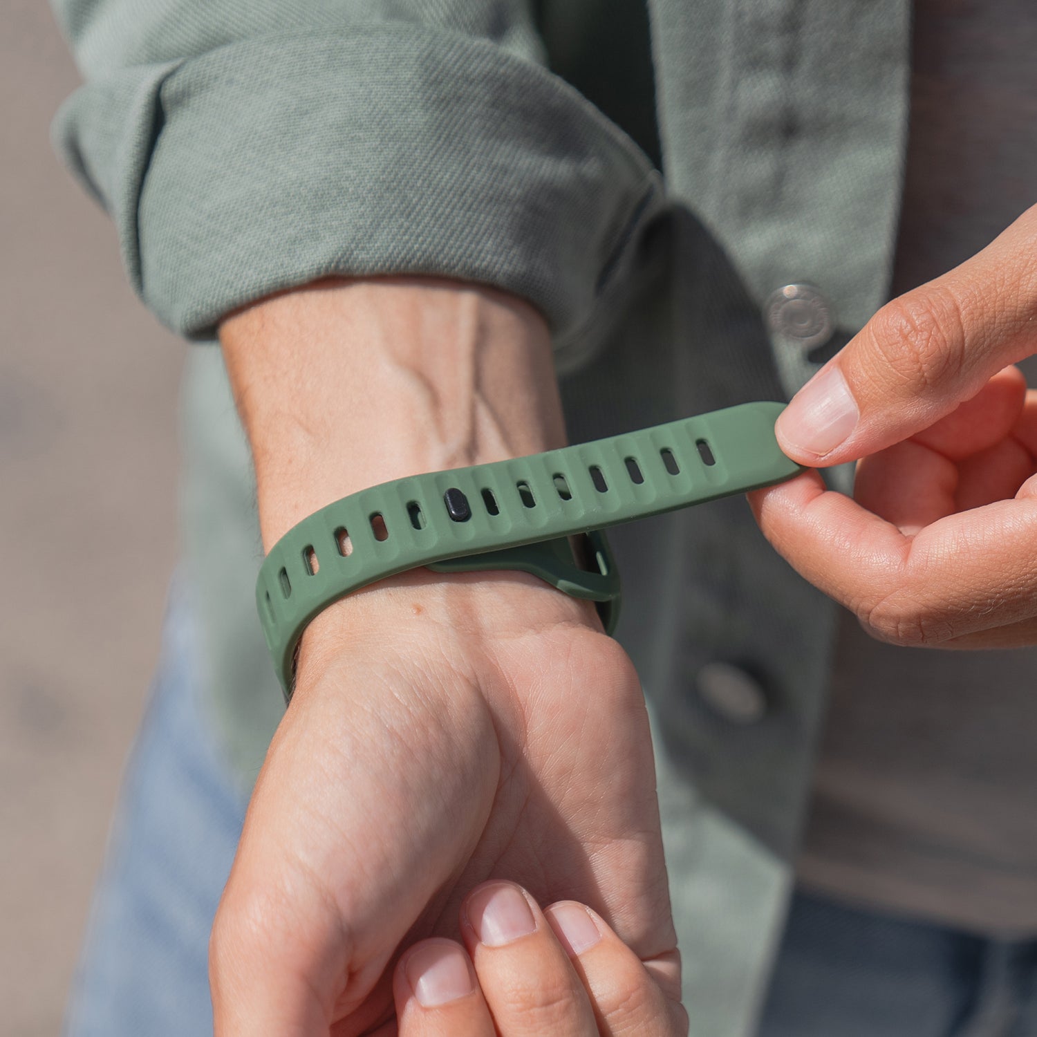 Eine Person befestigt ein grünes arktisband Apple Watch Silikonarmband "Active" von arktisband an ihrem linken Handgelenk. Das atmungsaktive Design bietet Tragekomfort für den Alltag und passt gut zu ihrer grünen Jacke und Jeans.