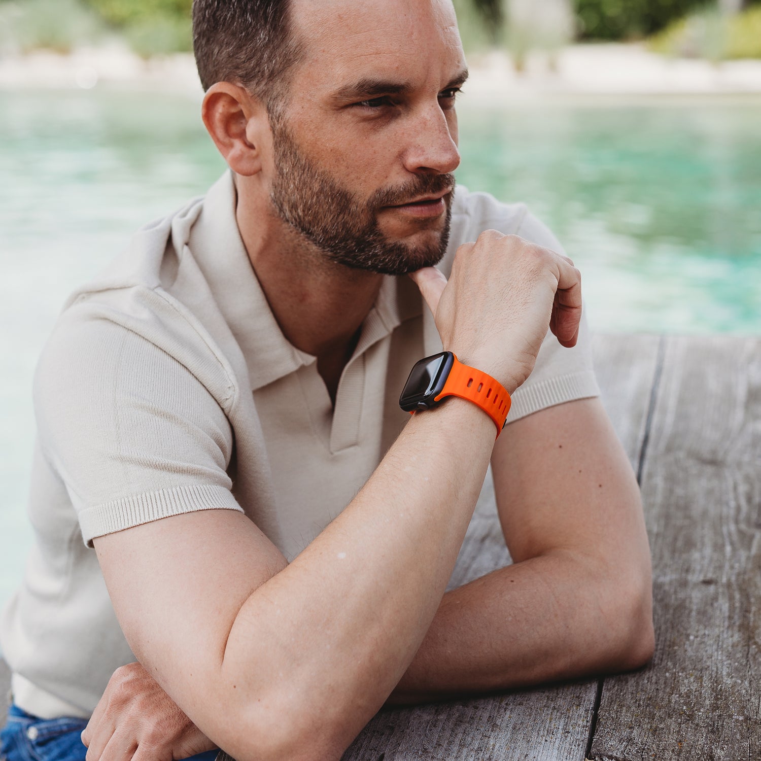 Ein Mann mit kurzen Haaren und Bart sitzt an einem Pool. Er trägt ein beiges Kurzarm-Poloshirt und das arktisband Apple Watch Silikonarmband „Active“ mit atmungsaktivem Design und orangefarbenem Silikonband.
