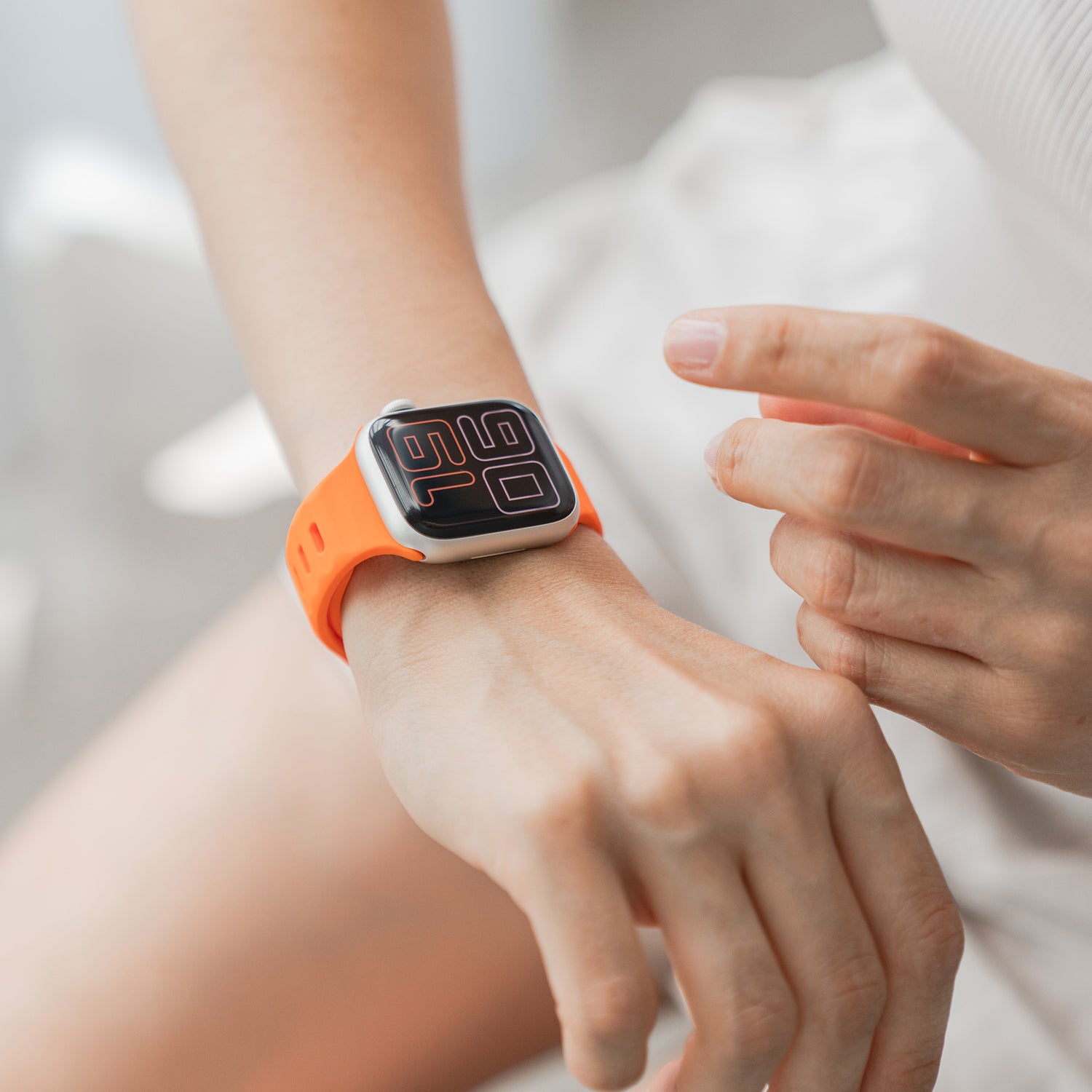 Eine Person in weißer Kleidung berührt ihr arktisband Apple Watch Silikonarmband „Active“ (orange) von arktisband, mit großem Digitaldisplay und atmungsaktivem Design, vor unscharfem Hintergrund.