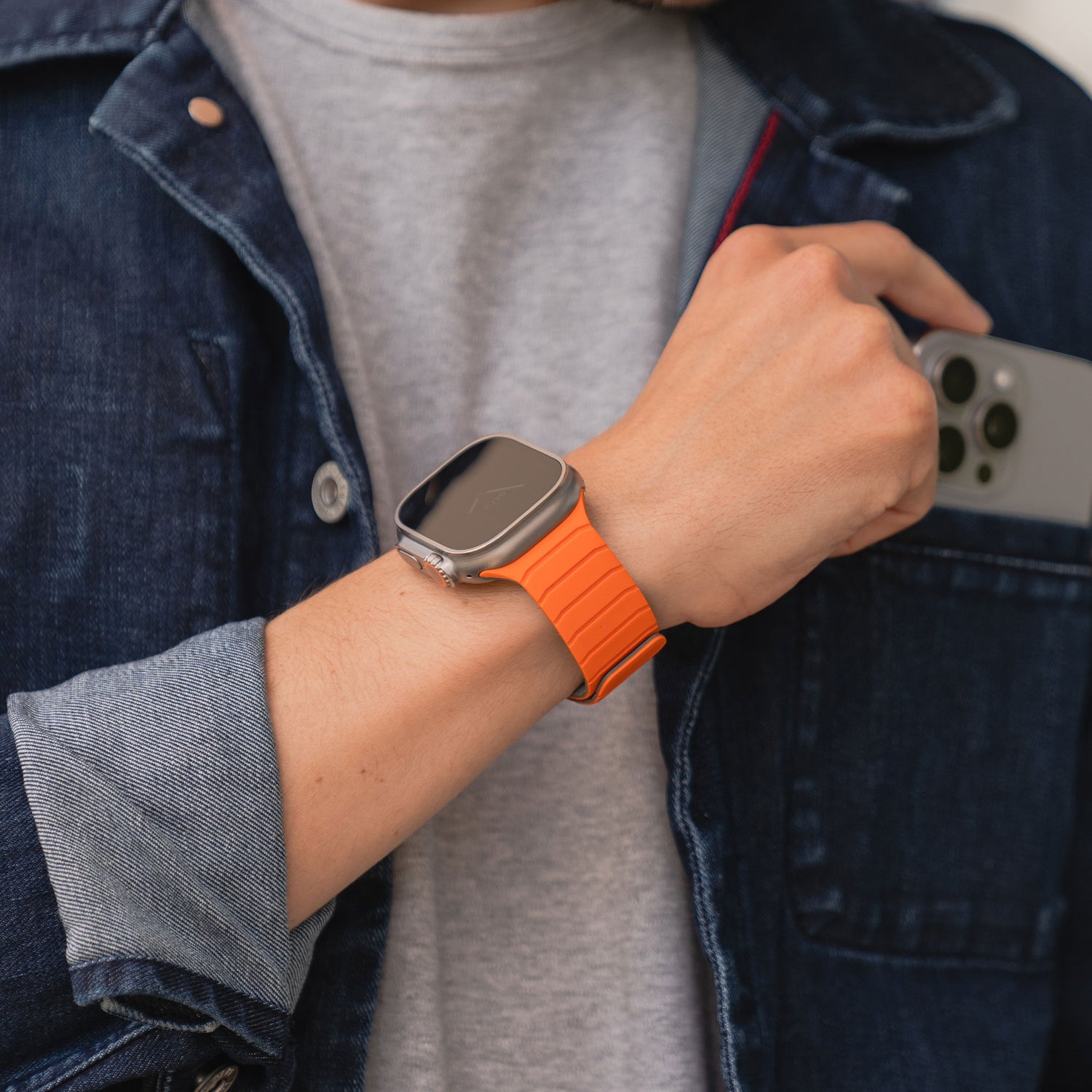 Eine Person in grauem Hemd und dunkler Jeansjacke hält ein Smartphone in der Hand und trägt das magnetische Armband „MagFlow“ der arktisband Apple Watch mit geschmeidigem Silikon und magnetischem Halt am Handgelenk.