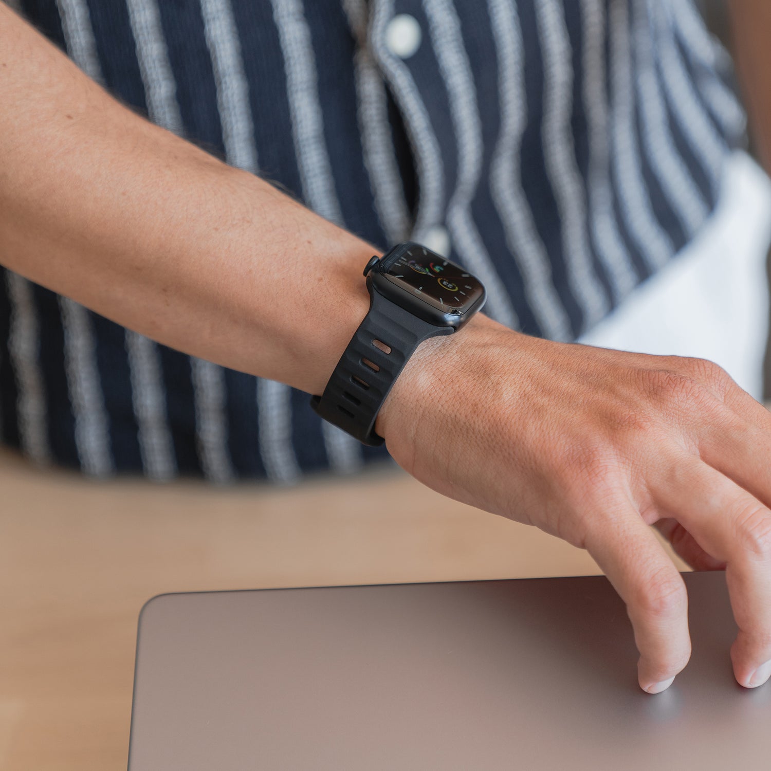 Eine Person trägt das schwarze arktisband Apple Watch Silikonarmband „Active“ mit atmungsaktivem Design und legt ihre Hand auf einen geschlossenen silbernen Laptop. Sie trägt ein kurzärmeliges, marineblau-weiß gestreiftes Hemd.