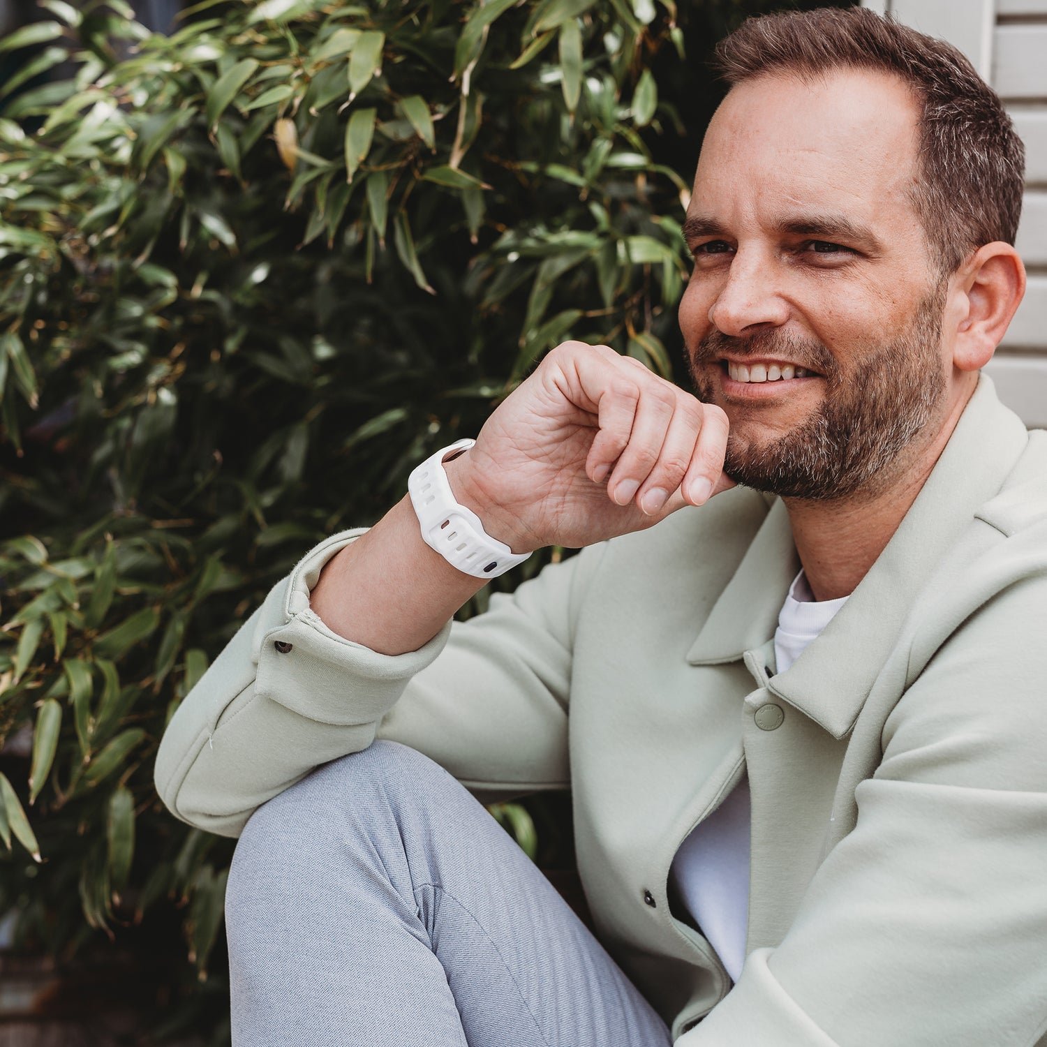 Ein Mann mit kurzen braunen Haaren und Bart lächelt draußen im Grünen. Er trägt eine hellgrüne Jacke, ein weißes Hemd, eine graue Hose und das arktisband Apple Watch Silikonarmband „Active“ von arktisband, seine Hand liegt vor seinem Gesicht.