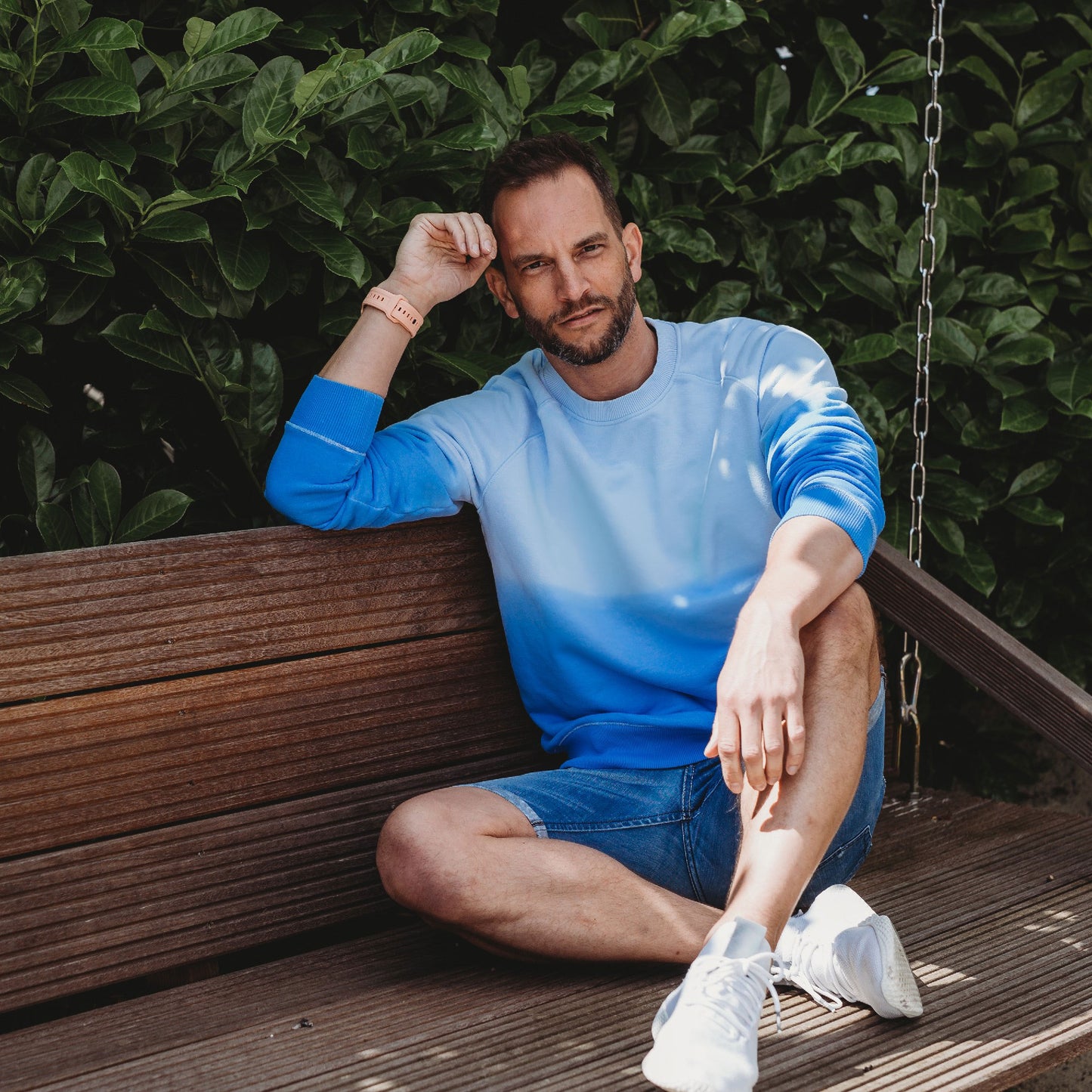 Ein Mann mit kurzen Haaren und Bart sitzt im Freien auf einer Holzbank. Er trägt ein blaues Sweatshirt, Jeansshorts, weiße Turnschuhe und ein arktisband Apple Watch FKM Sportarmband „Sweat“ von arktisband. Im Hintergrund sind grünes Laub und ein Teil einer Schaukelkette zu sehen.
