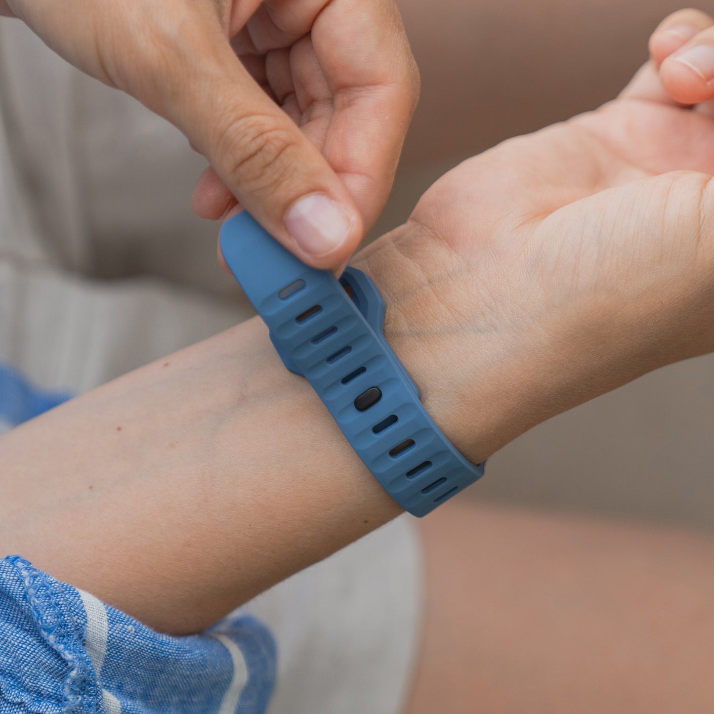 Eine Person befestigt mit der rechten Hand ein Arktisband Apple Watch Silikonarmband „Active“ in Blau am linken Handgelenk. Das Band bietet angenehme Atmungsaktivität. Sie trägt ein blau-weiß gestreiftes Kleidungsstück.