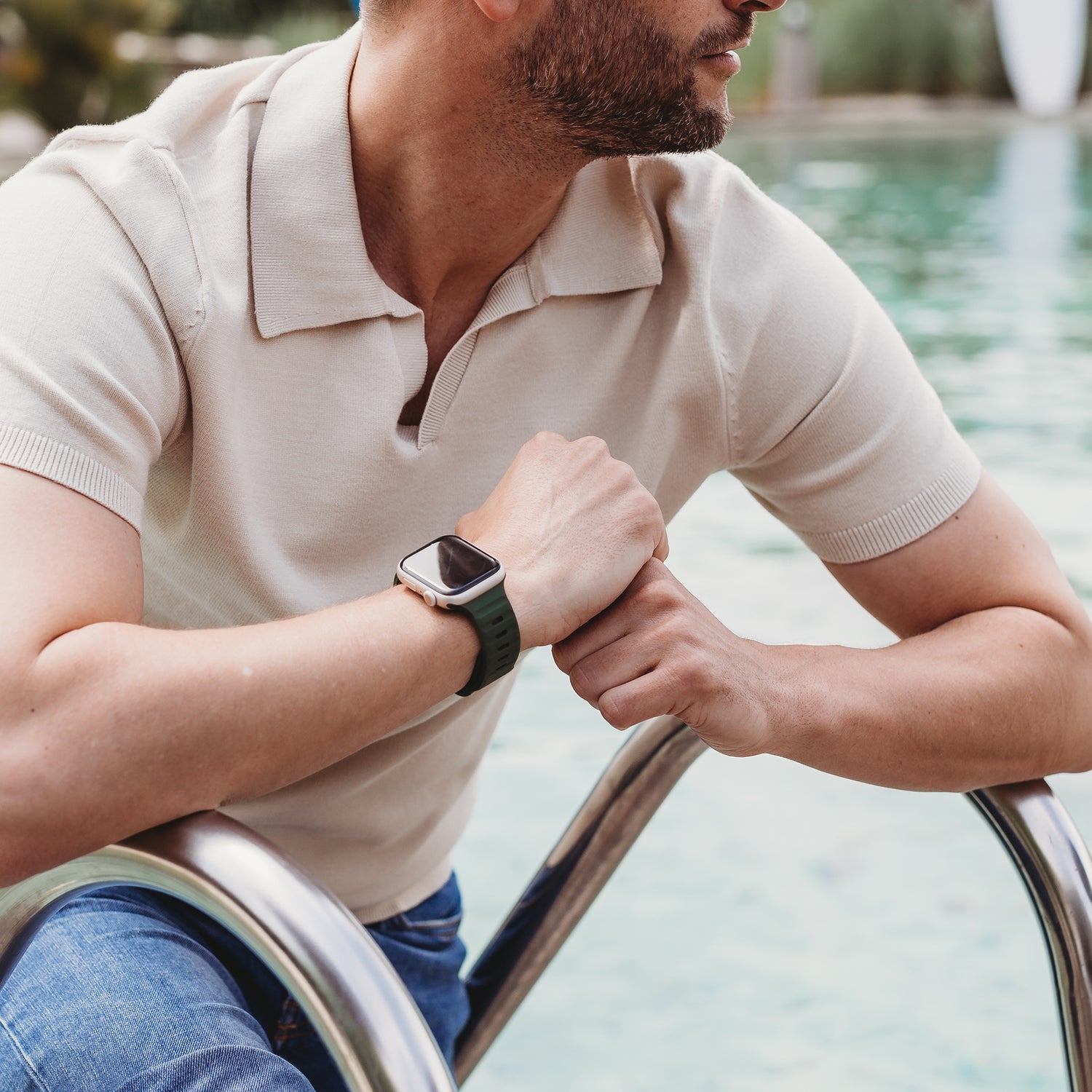 Ein Mann in einem kurzärmeligen beigen Poloshirt und Jeans sitzt auf den Stufen am Pool und blickt zur Seite. An seinem linken Handgelenk trägt er das schwarze Arktisband Apple Watch Silikonarmband „Active“. Hinter ihm ist das glitzernde Poolwasser zu sehen.